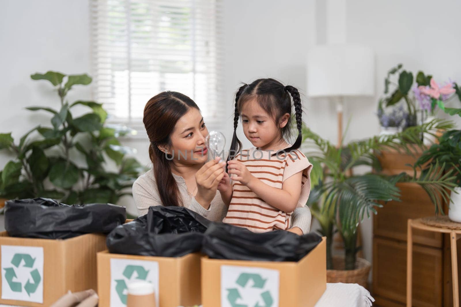 A caring mother shows her daughter a lightbulb, explaining the importance of recycling as they sort materials at home, promoting environmental awareness.