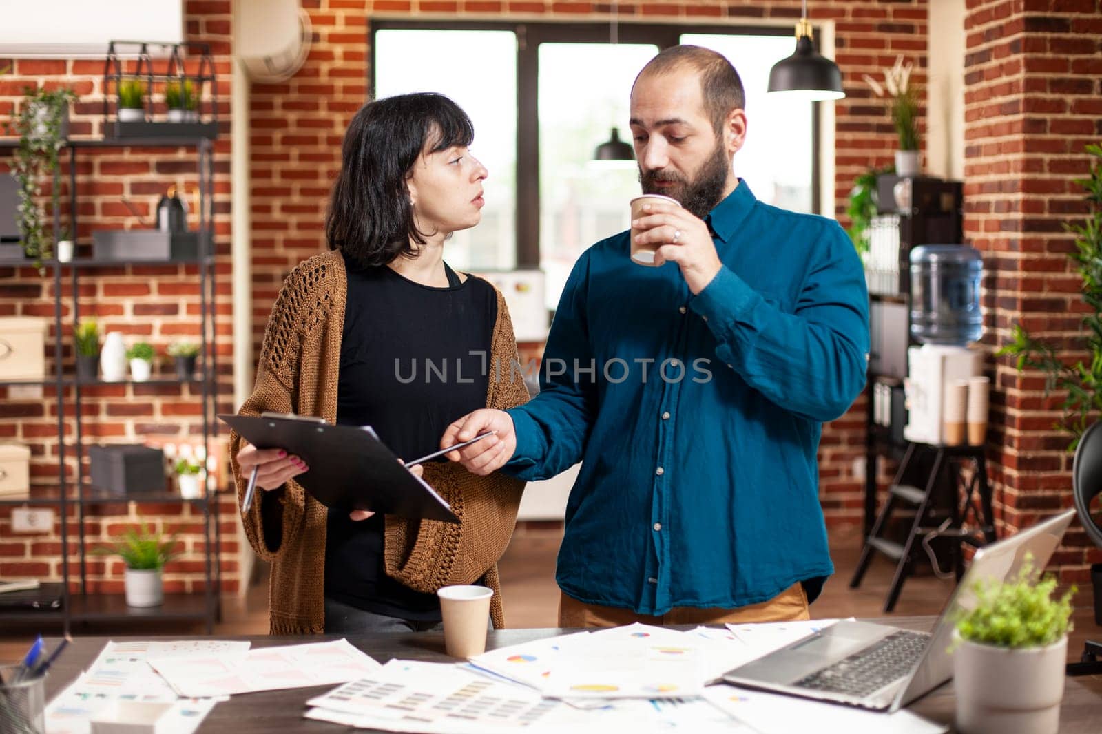 Businessman drinking coffee and assisting his female coworker with business analytics of startup company. Young woman with clipboard, showing strategy ideas to manager with beard at office desk.