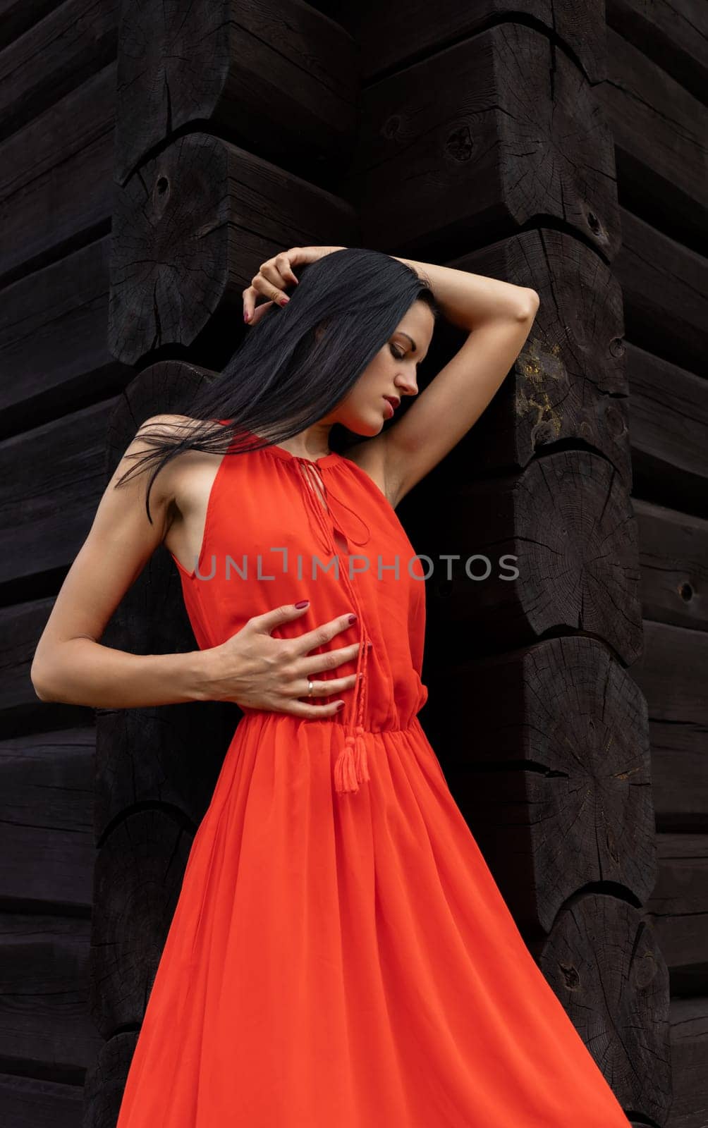 A slender woman in a vibrant red dress shields her eyes, gazing towards the horizon against a rustic, dark wooden wall.