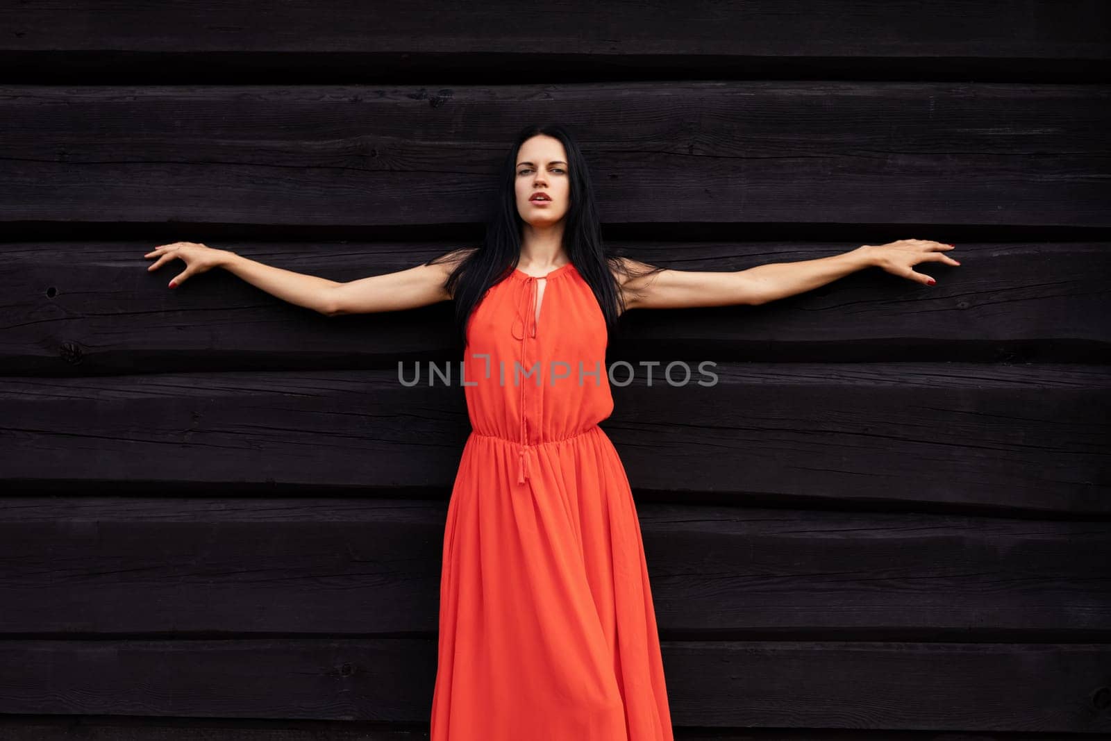 A slender woman in a vibrant red dress shields her eyes, gazing towards the horizon against a rustic, dark wooden wall.