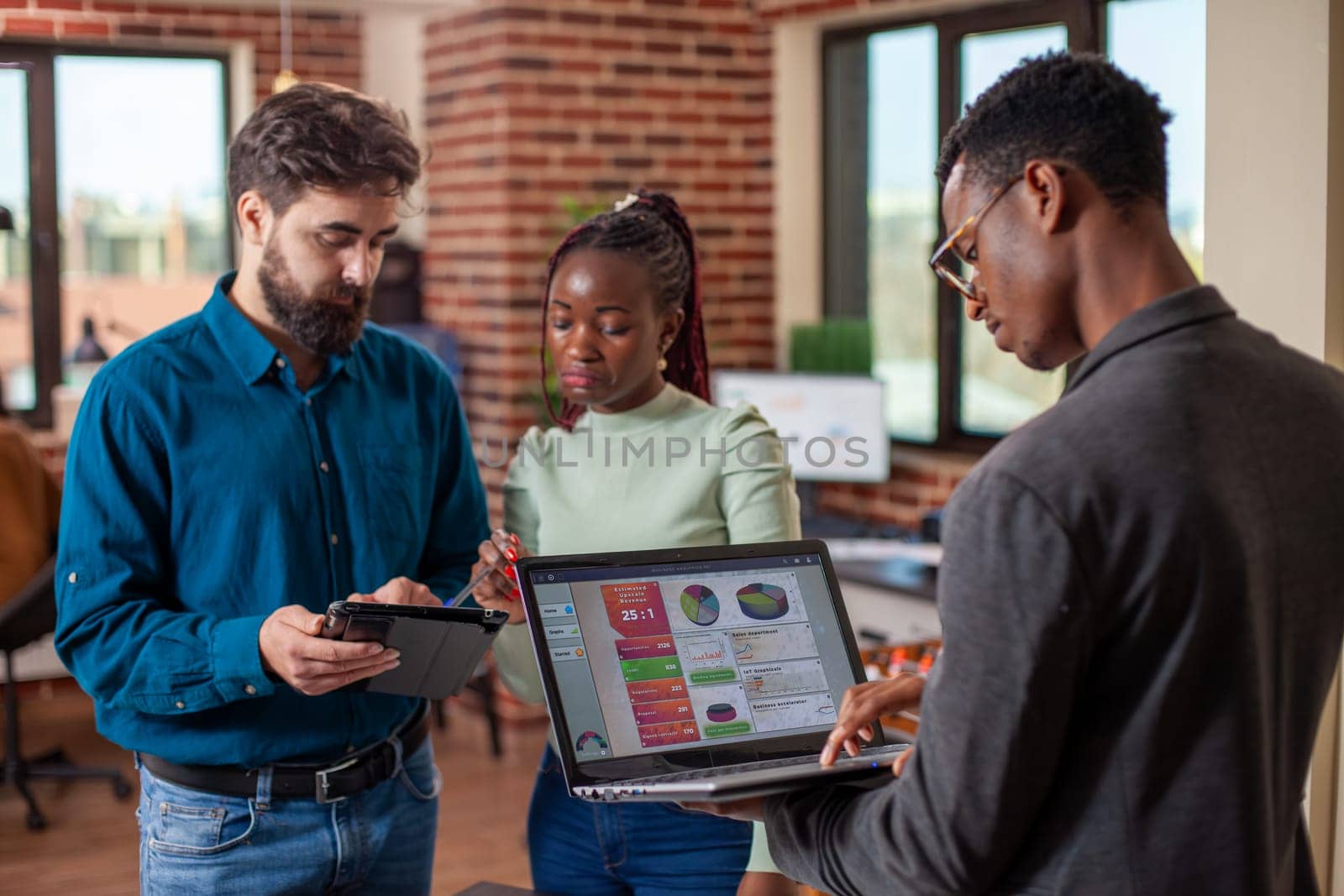 Colleagues utilizing digital technology during their discussion in modern workspace. Black man using laptop to retrieve marketing data as his male and female coworkers review strategy plans on tablet.