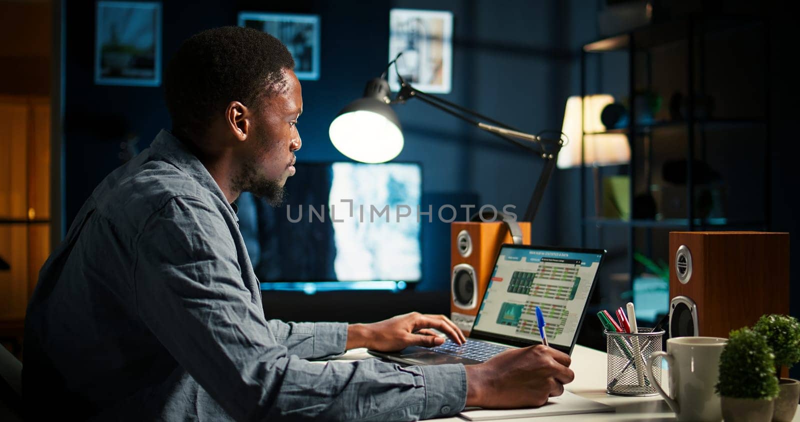 Self employed Black man multitasking at home taking handwritten notes and browsing the web for project research. Managing schedule and tasks with a planner to boost productivity. Camera A.