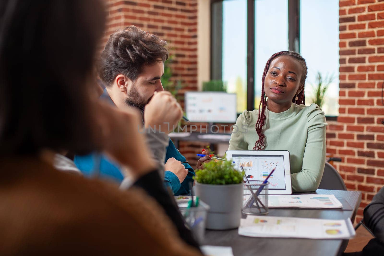 Black businesswoman with tablet, presents market strategy ideas to her employees in modern workspace. Female entrepreneur seated at table with her startup team, using device to describe company plans.