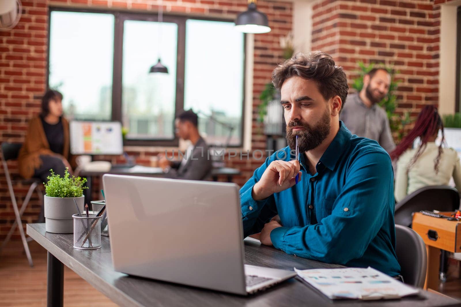 Male researcher studying documents with pen in hand, focused on project development at desk. Caucasian employee seated in brick wall office, tapping his chin and reviewing business plans on laptop.