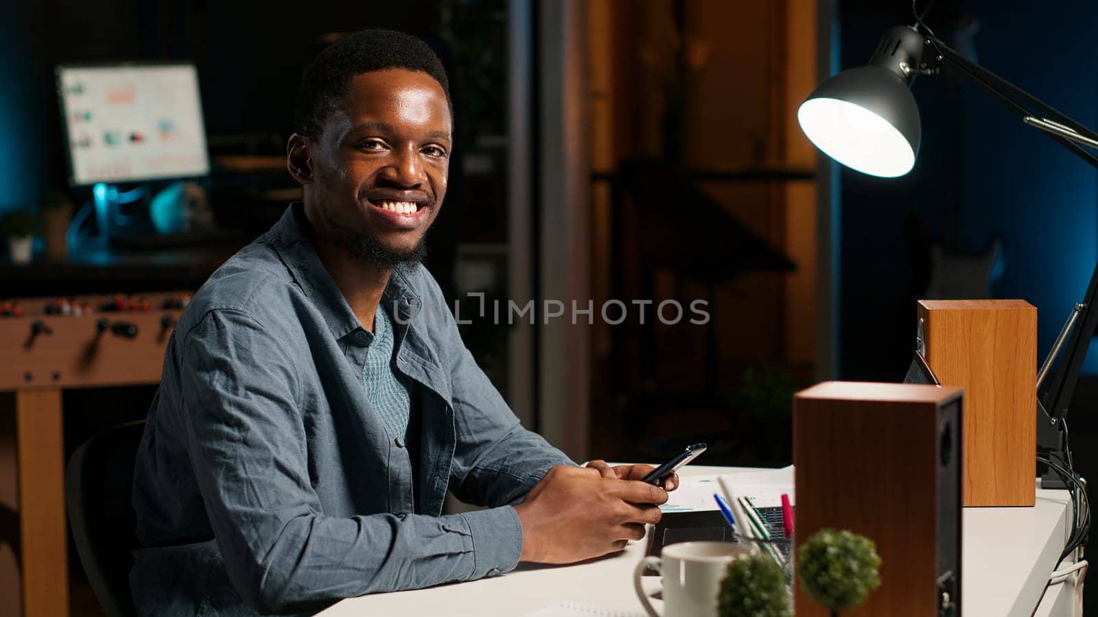 Portrait of african american guy scrolling on his mobile phone at home, takes break from his freelance tasks and online work. Young adult texting messages on social media app, relaxation. Camera B.