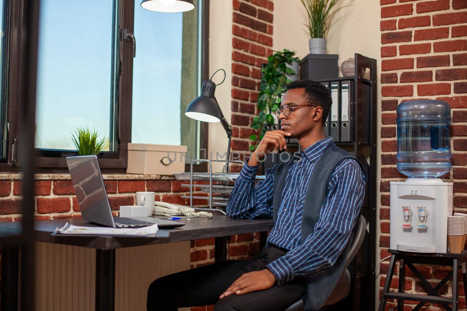 Young businessman sitting at office desk with wireless earphones, watching online presentation on his personal computer. Black male employee in brick wall workplace, attends virtual training on laptop