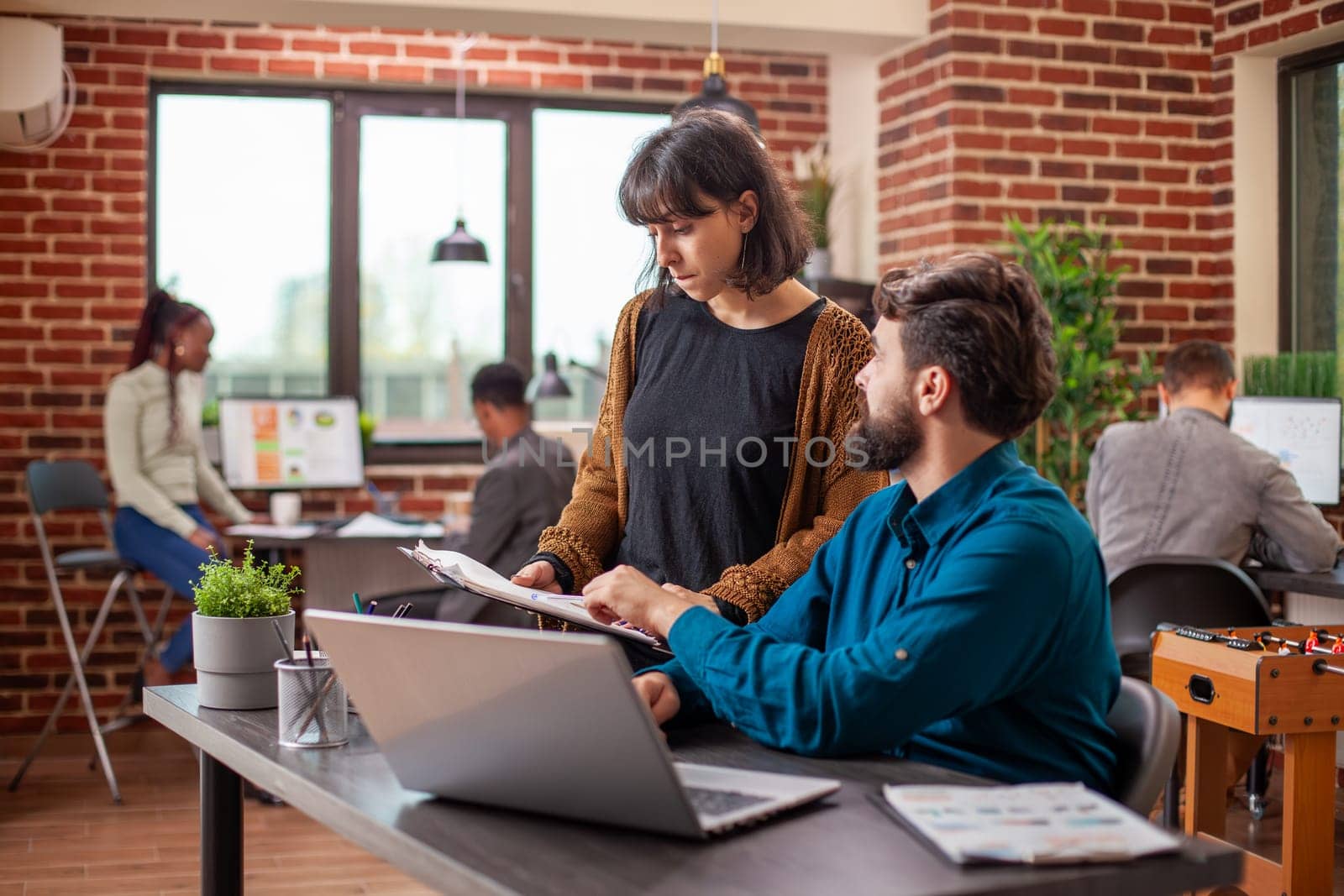 Male manager collaborates with female employee on business plan using documents and online data on laptop. Businesswoman holds clipboard and shows company research statistics to coworker in office.