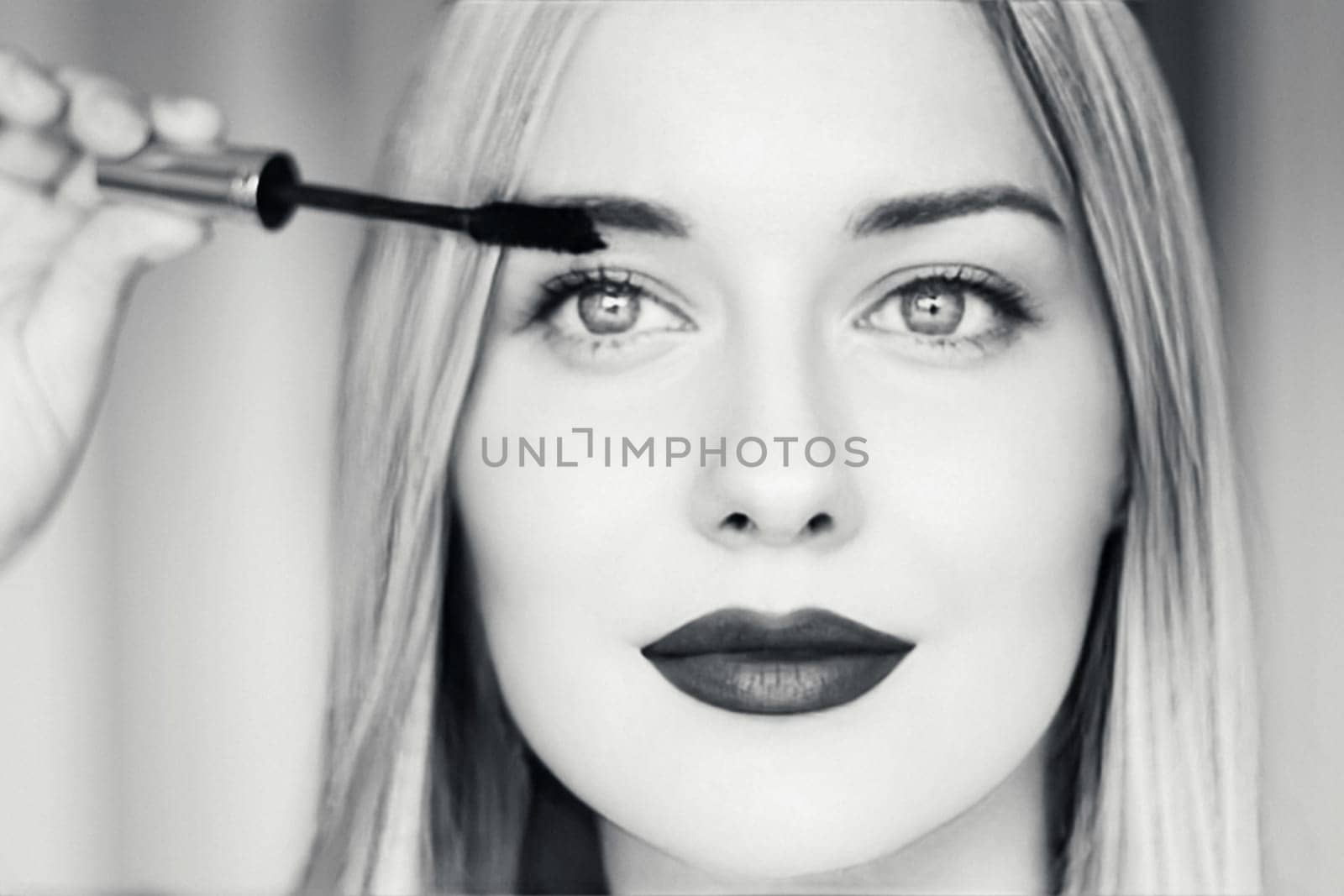 Close-up of a beautiful blonde woman applying mascara, with dark lipstick, bright eyes, and long straight hair. Black and white portrait for beauty, makeup, fashion, or cosmetic themes