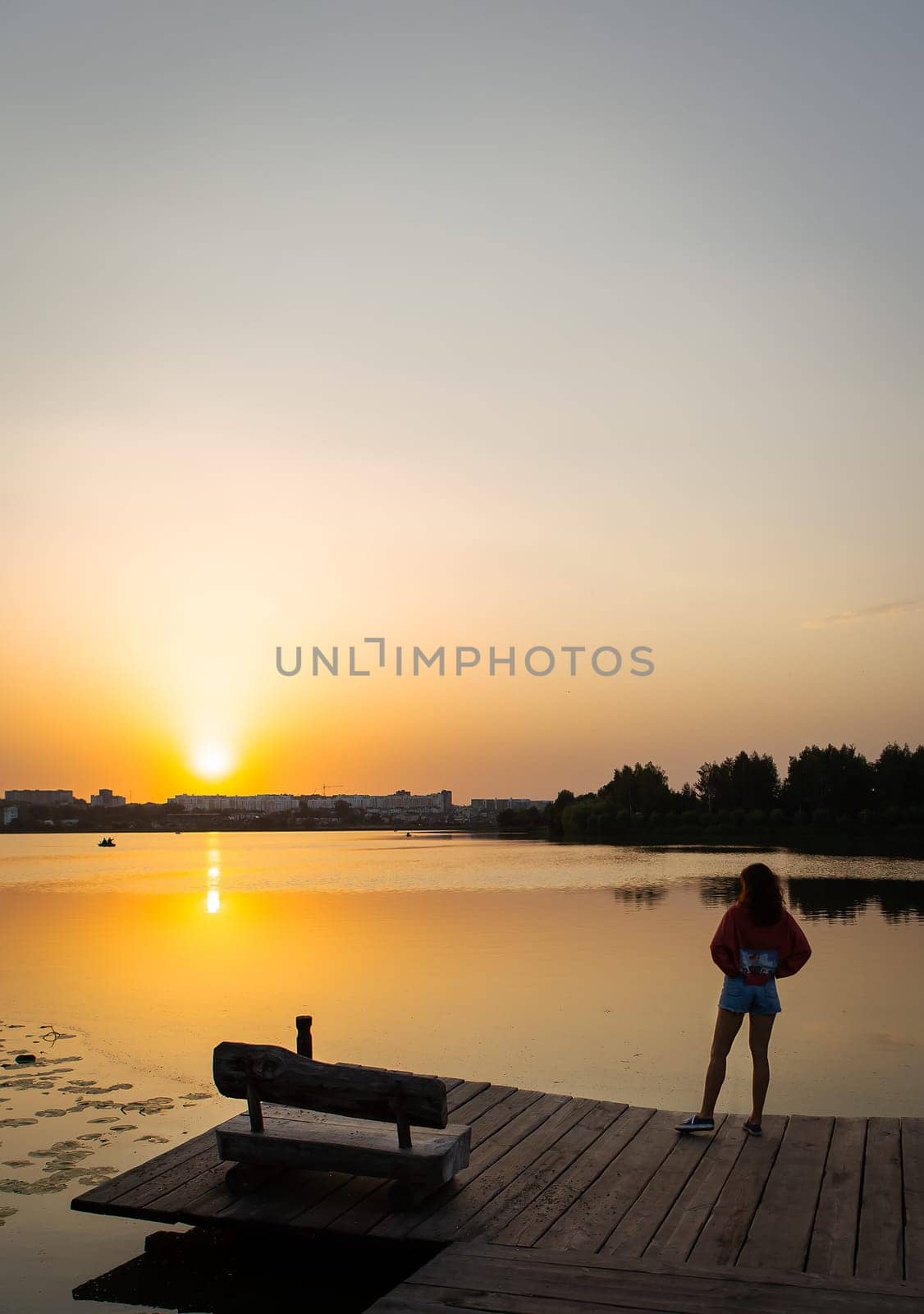 A woman standing on a wooden dock, gazing at a serene sunset over a calm lake with reflections and a tranquil atmosphere.