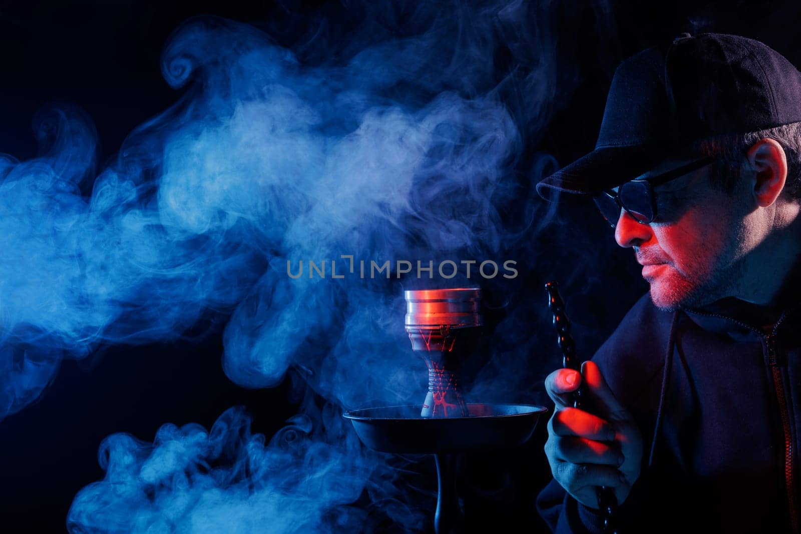 Man smoking hookah, adjusting hot charcoal with tongs in a smoky, moody atmosphere, illuminated by red and blue lighting,