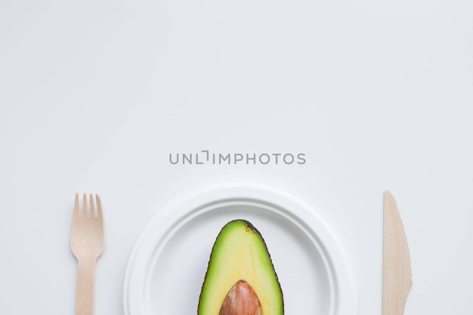 Minimalistic half avocado on white plate with wooden cutlery.