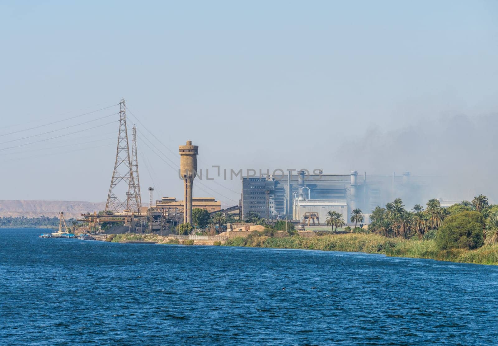 A view of the Edfu steel factory, including industrial buildings, smokestacks, and power lines, seen from the Nile River