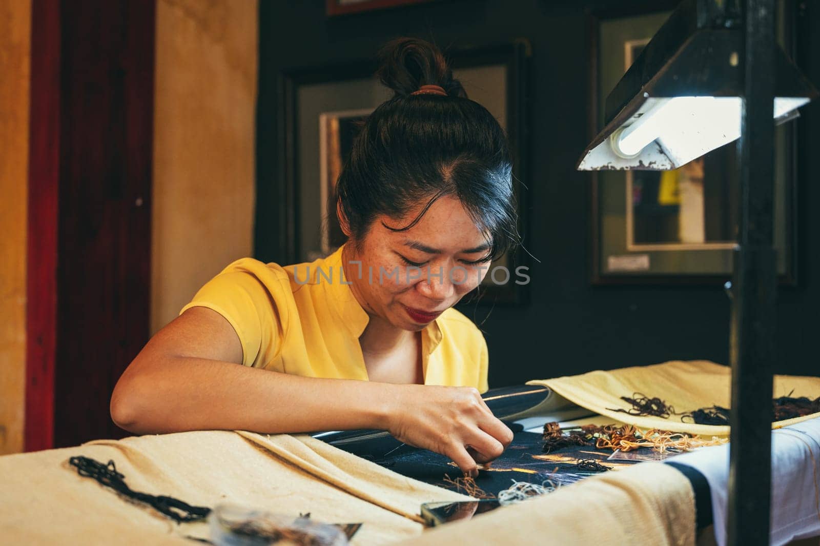 Happy Vietnamese female embroiderer embroiders a silk painting in a workshop in the old town in Hoi An city in Asia. Hoi An, Vietnam - December 1, 2024
