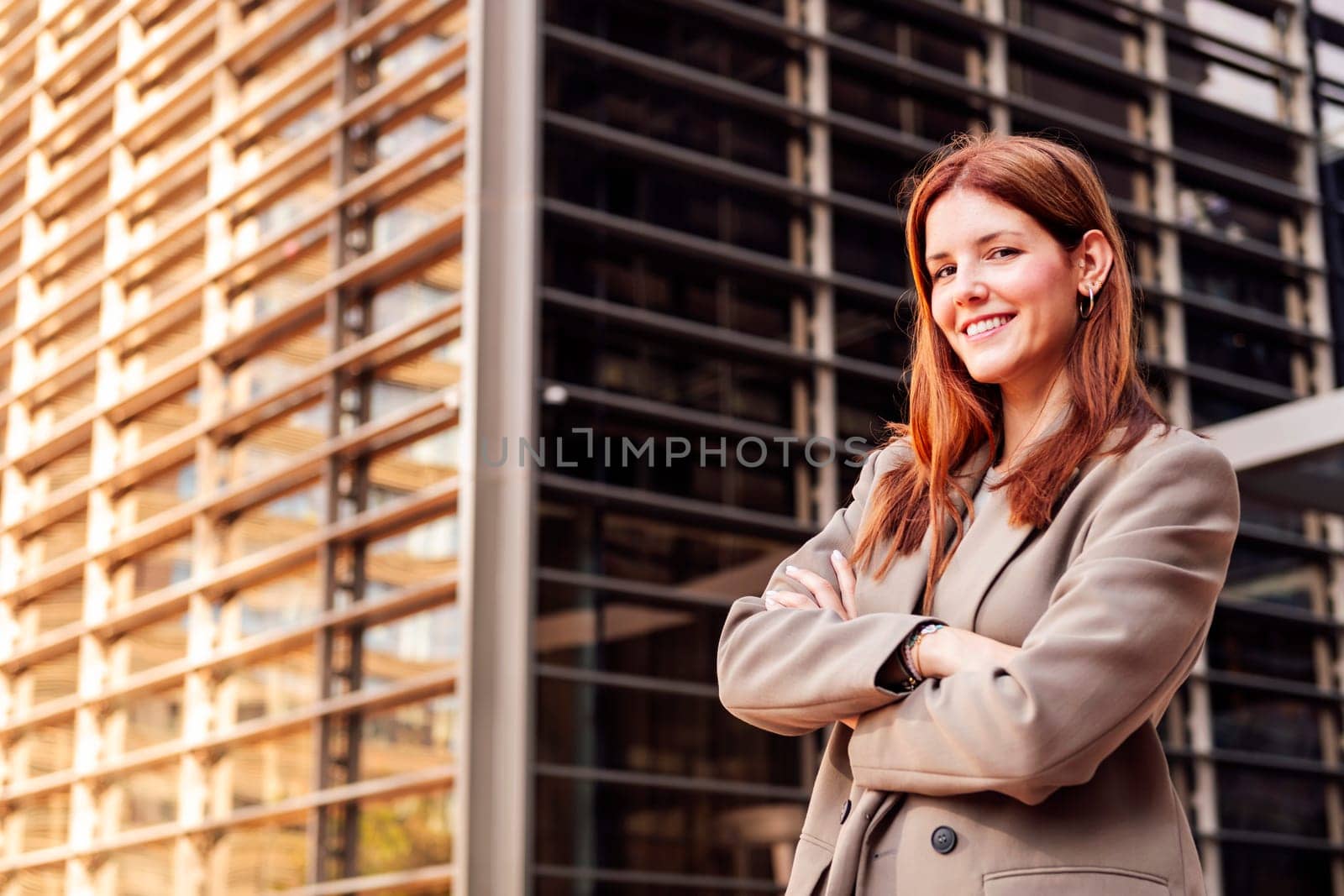 young adult woman in suit jacket standing looking at camera smiling happy with arms crossed in front of an office building, concept of entrepreneur and business, copyspace for text