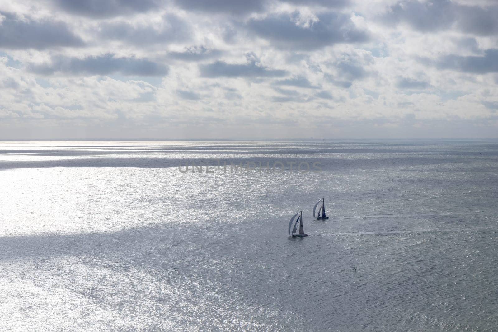 Aerial view of two sailboats plowing together in calm sea. Nearby, kite surfers soar with colorful sails, carving waves and leaping into the air