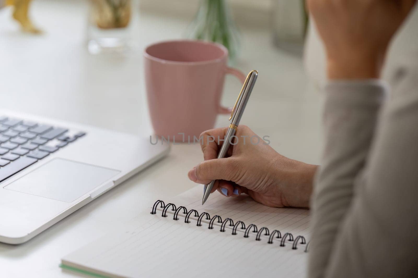 Focusing intently, a woman writes in her notebook next to a laptop, demonstrating the balance of writing and digital work while sipping coffee.