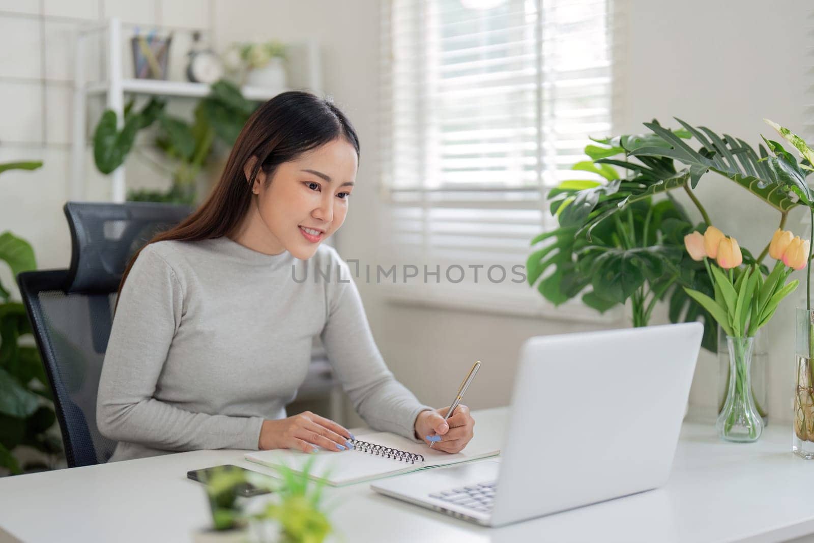 A young woman actively participates in a virtual meeting, jotting down key points in her notebook amidst a bright, plant-filled workspace.