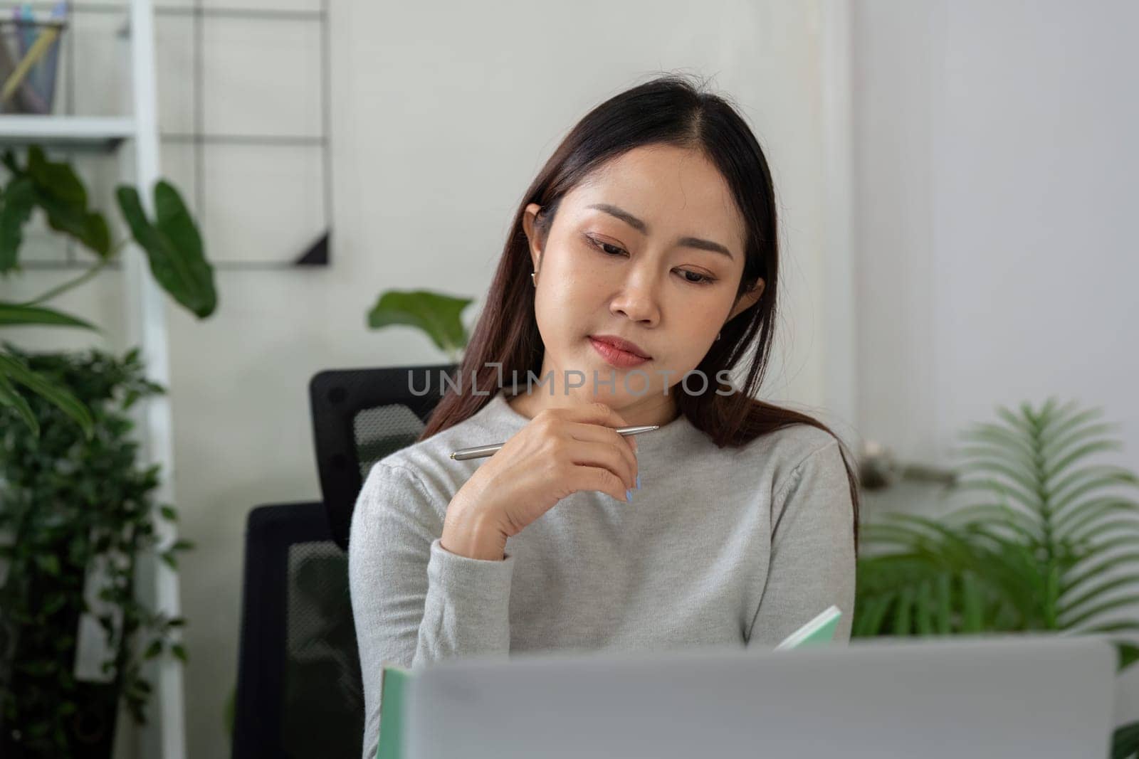 The woman reflects on her work while holding a pen and jotting down ideas in her notepad. She embodies the essence of balance and diligence in remote working.
