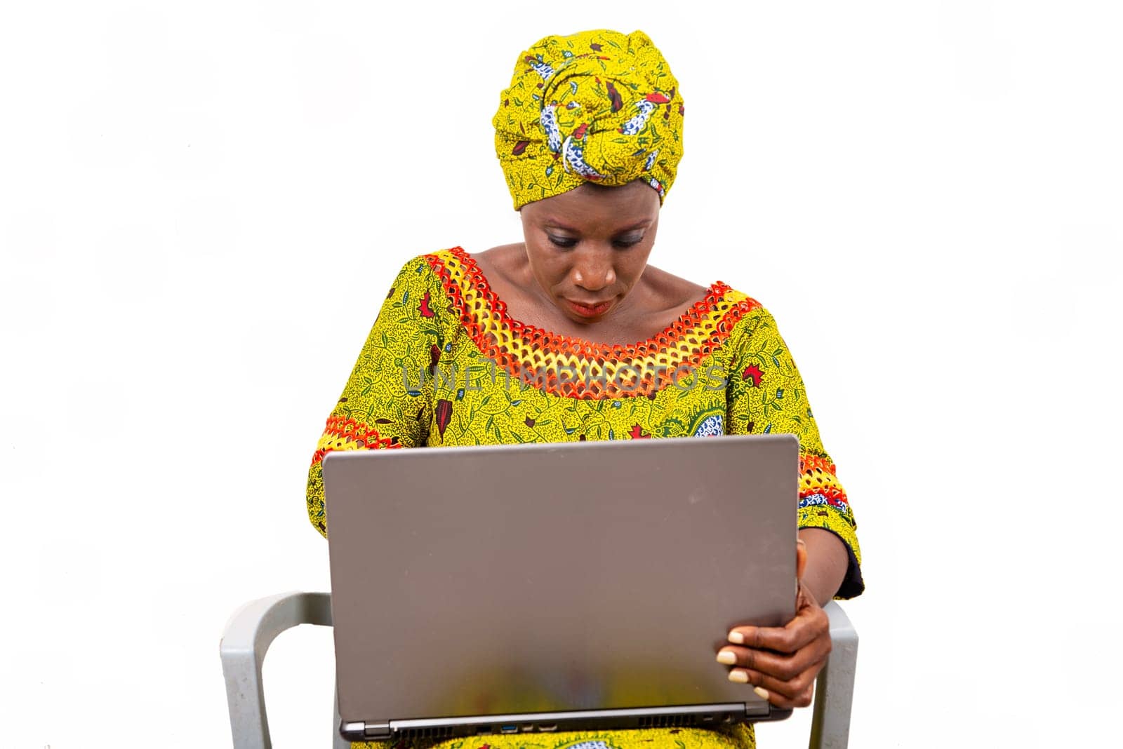 Beautiful African adult woman wearing a loincloth and a green scarf on her head is sitting on a chair manipulating her computer