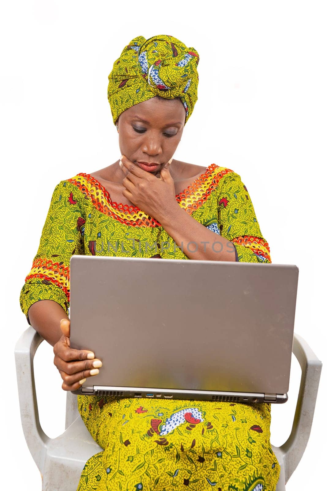 Beautiful African adult woman wearing a loincloth and a green scarf on her head, sits on a chair with her computer thinking about putting her hand on her chin