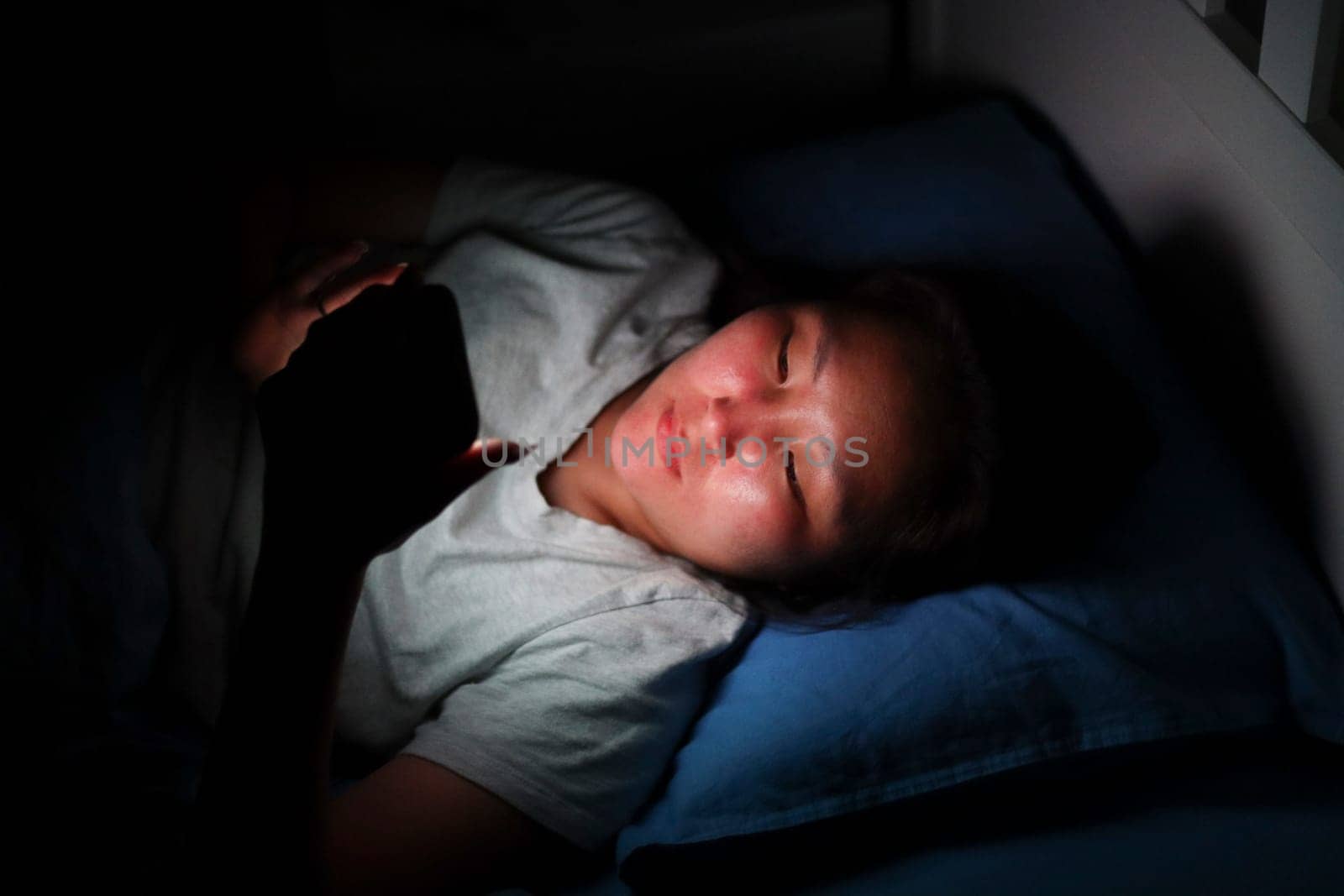 Asian woman lying in bed using smartphone at night, illuminated by screen light