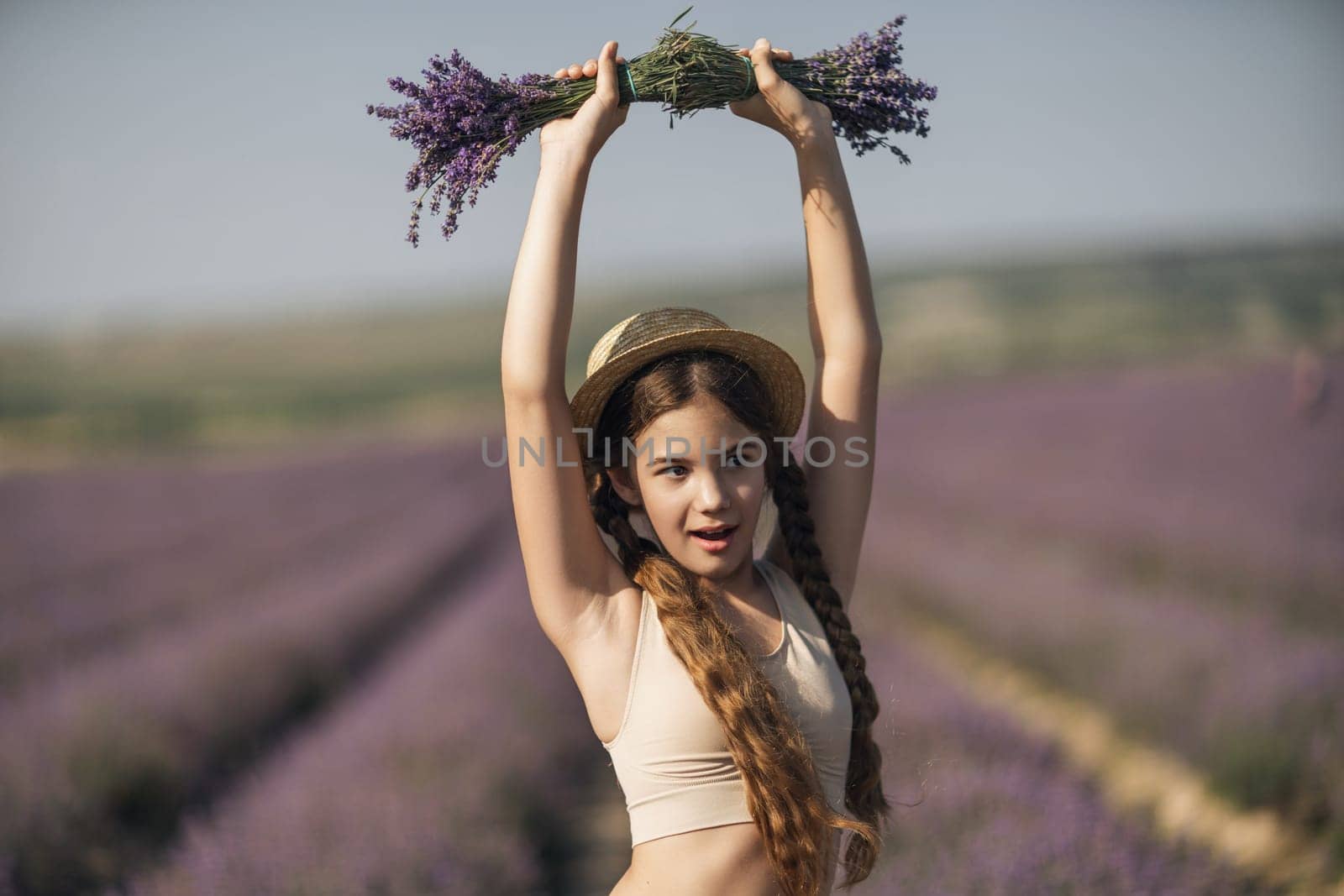 girl is holding a bunch of lavender purple flowers in her hands and wearing a straw hat. She is smiling and she is enjoying herself. The scene is set in a field of lavender, which adds to the peaceful
