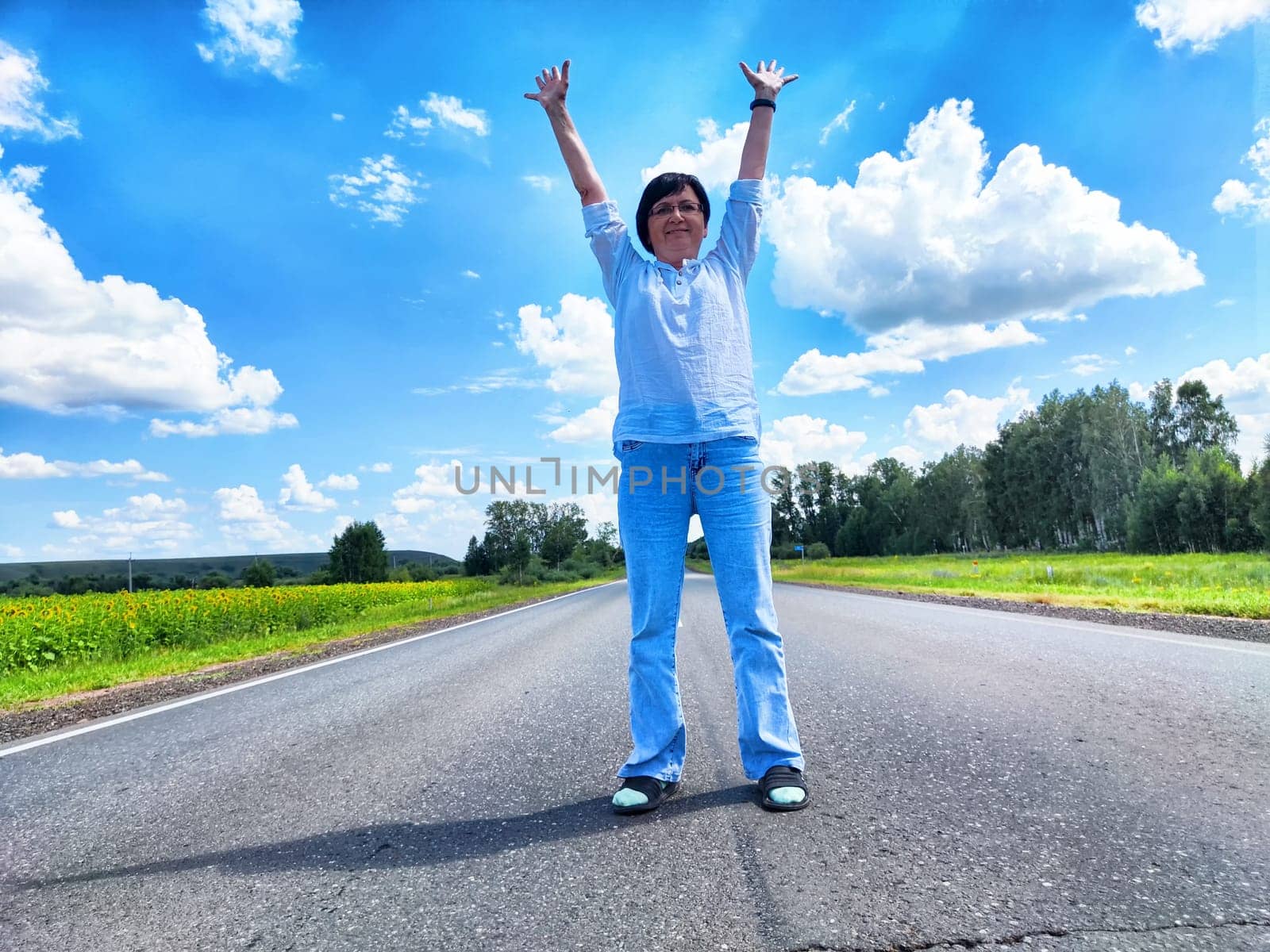 Middle-aged blogger celebrates her journey by stretching arms wide on a picturesque country road under a bright blue sky.