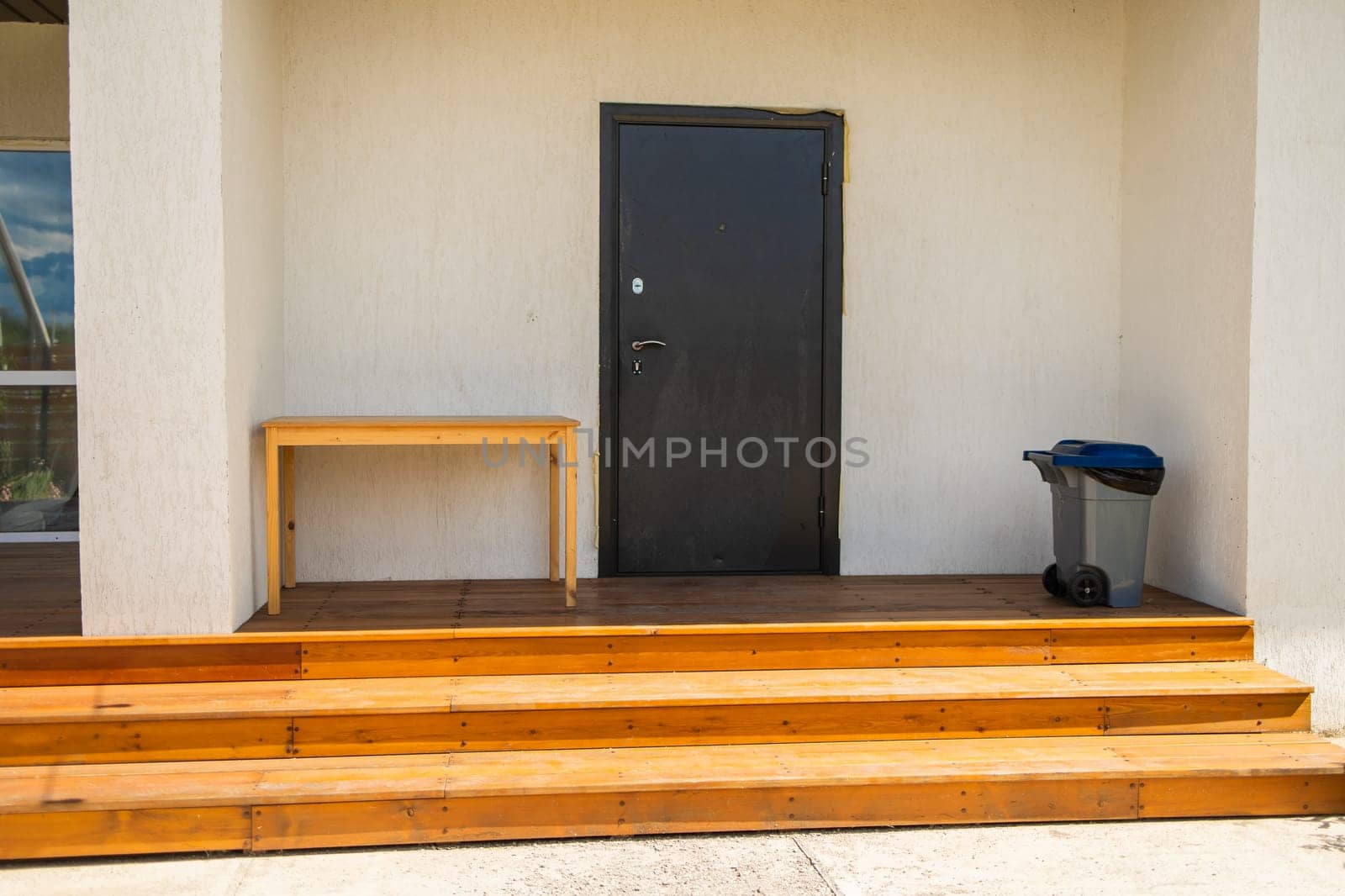 Black entrance door with trash bin and wooden table. Neat home access and minimalistic outdoor arrangement for everyday use.