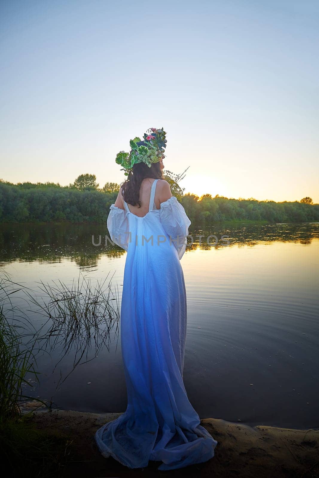 Slavic plump chubby girl in long white dress on the feast of Ivan Kupala with flowers and water in a river or lake on summer evening