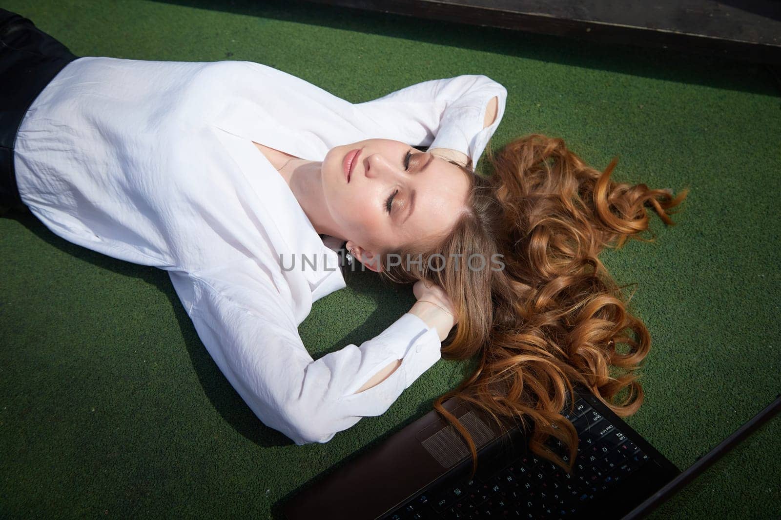A young woman with long curly hair enjoys a moment of relaxation on green grass, surrounded by sunshine and a laptop.