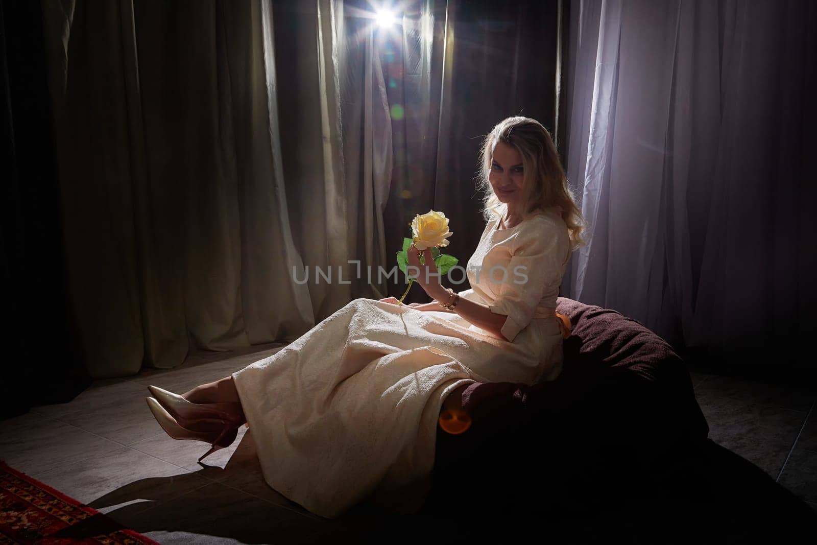 Woman with blonde hair enjoys a serene moment while modeling with a flower in a softly lit indoor space.