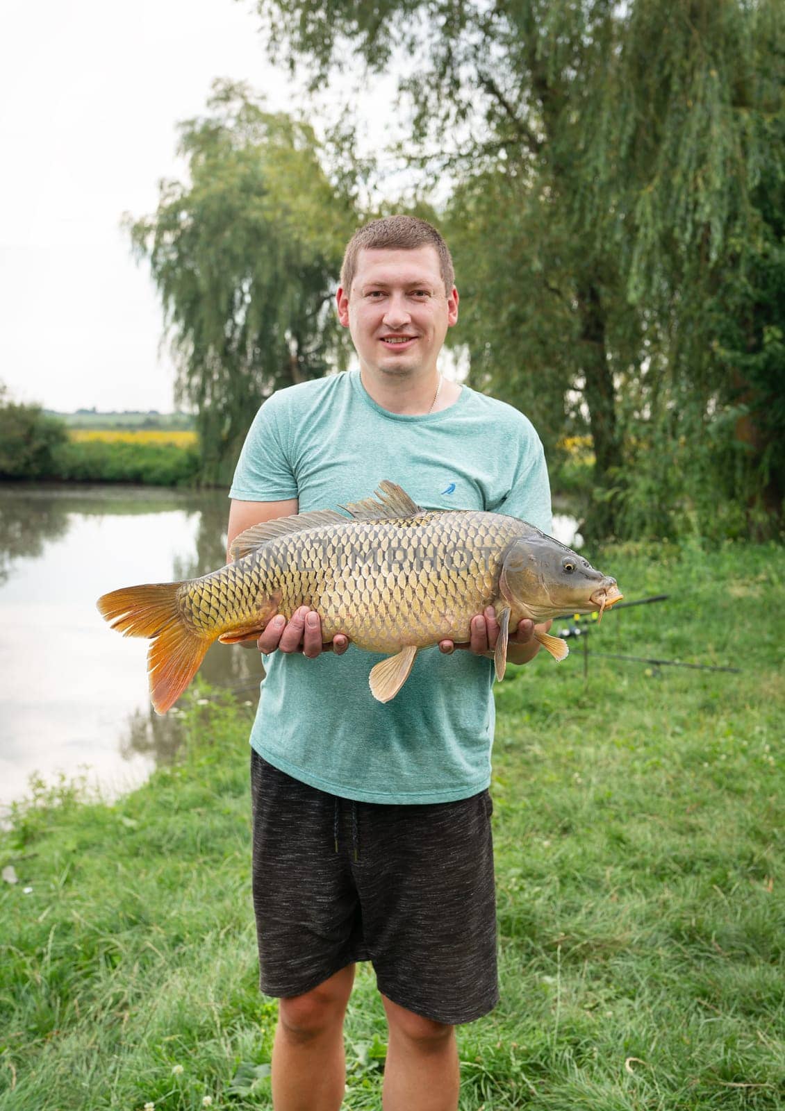 A fisherman proudly holding a large carp near a scenic lake, surrounded by lush greenery and a calm outdoor setting.
