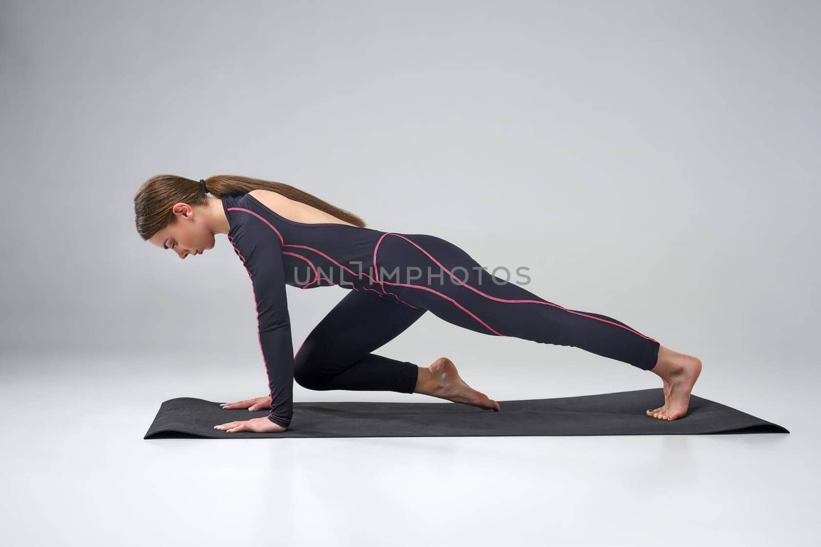 Flexible woman in black bodysuit exercising on yoga man in studio. Side view of concentrated brunette girl wearing tight sportswear, practicing yoga asana, isolated on grey. Concept of yoga.