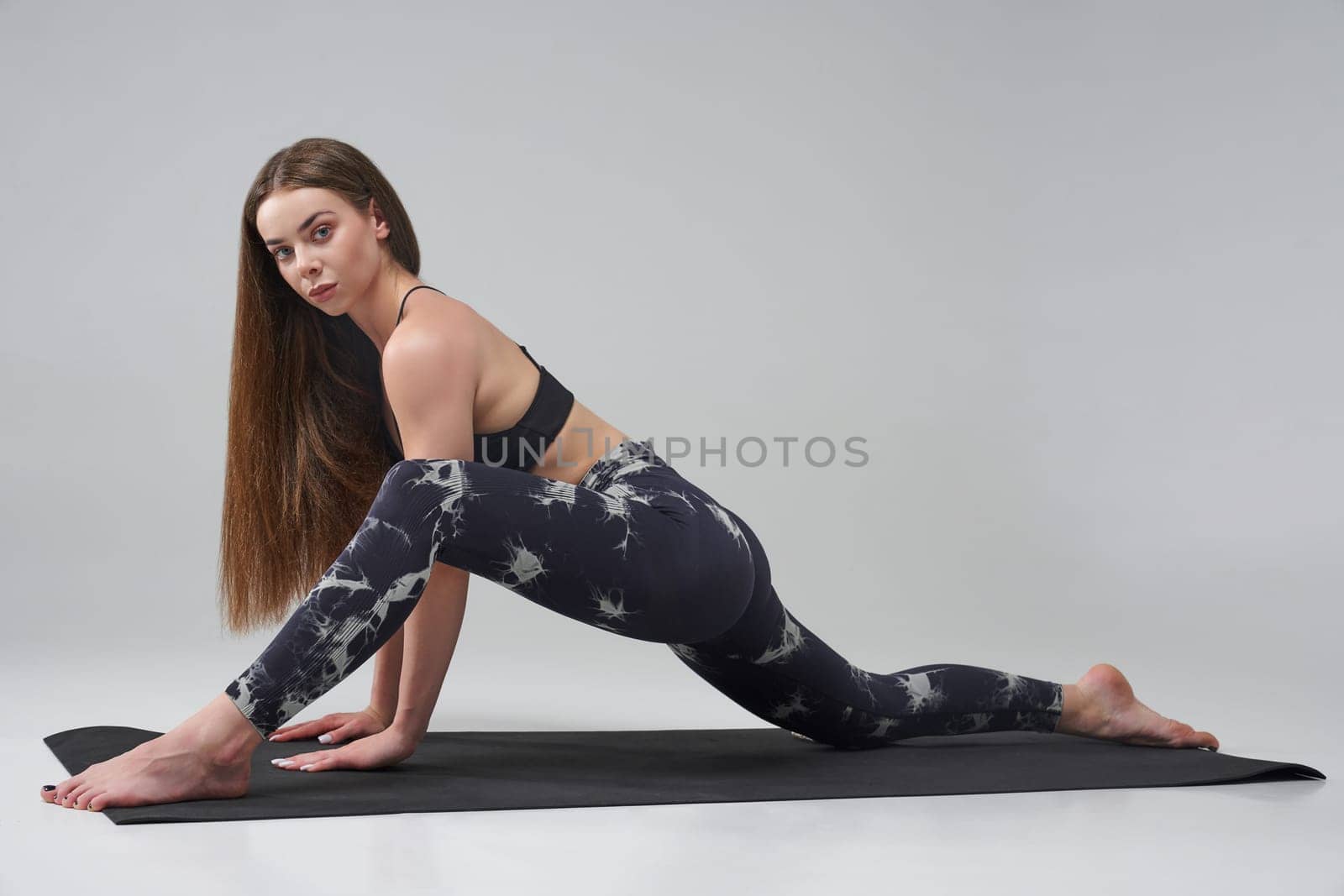 Barefoot Caucasian girl doing lizard position of yoga in studio. Side view of pretty female in activewear looking at camera, while practicing yoga on mat, isolated on grey background. Yoga concept.
