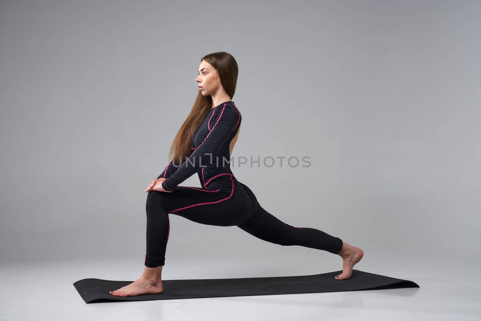 Slim woman in black bodysuit exercising on gym mat. Side view of calm Caucasian girl with long hair, practicing yoga, isolated on grey studio background. Concept of yoga, sport, healthy lifestyle.