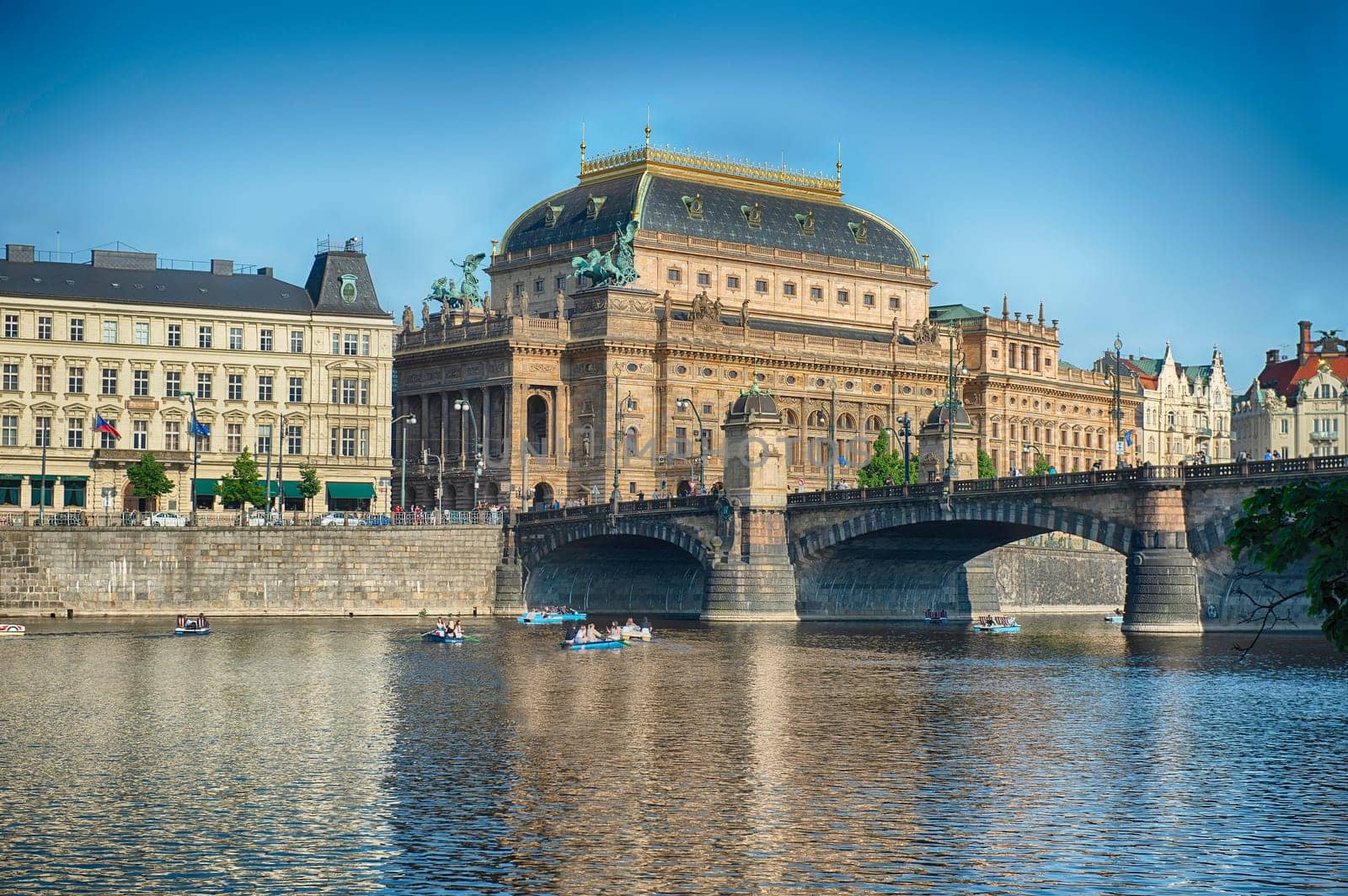 The National Theatre sitting on the bank of the Vltava river in Prague with tourists enjoying a sunny day on paddle boats