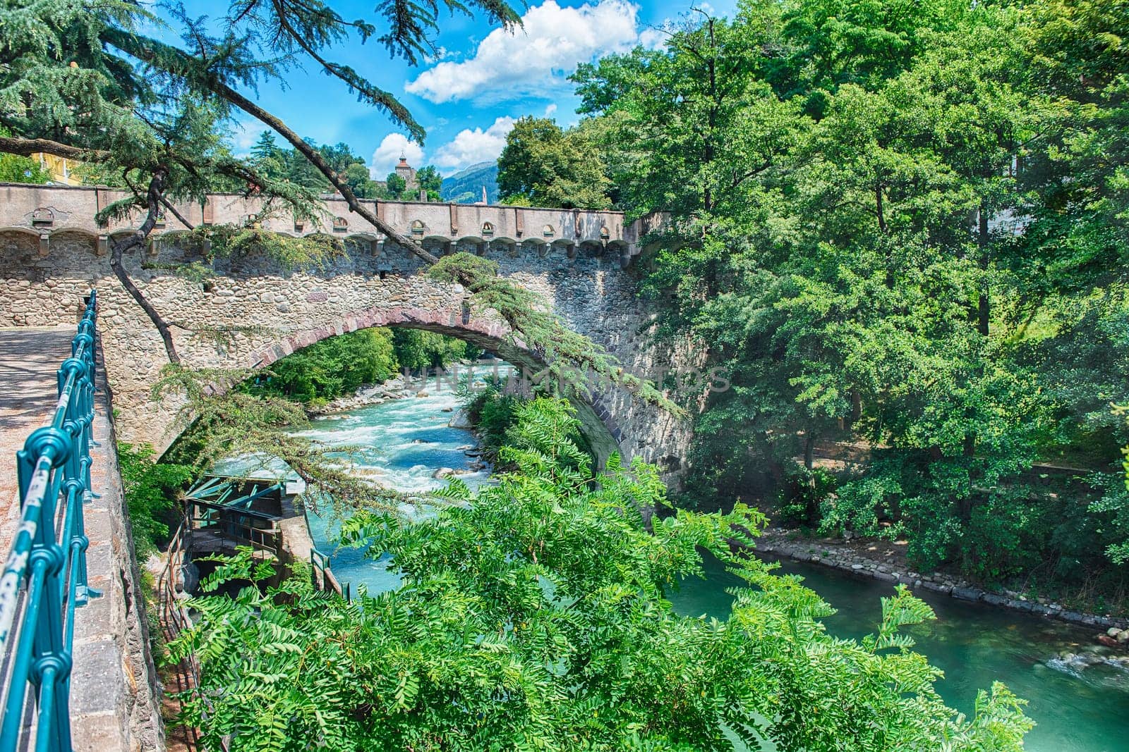 View of Steinerner Steg bridge crossing a river with lush green trees on both sides, iconic landmark in Merano, South Tyrol, Italy