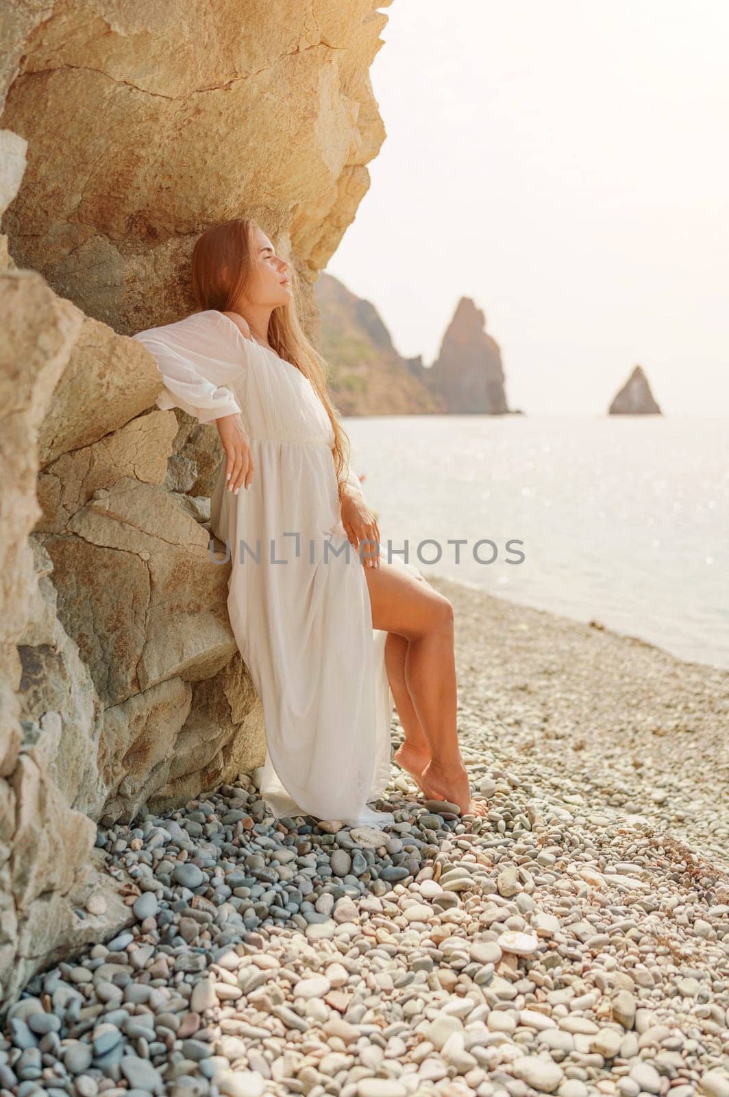 woman white dress standing on a beach, looking out at the ocean. She is lost in thought, with her hand on her hip. Concept of calm and introspection