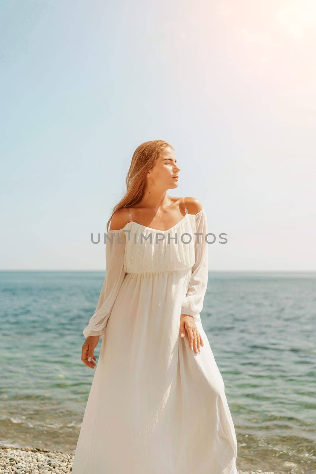 woman white dress stands on a beach, looking out at the ocean. She is in a contemplative or reflective mood, as she raises her hands above her head. Concept of peace and tranquility