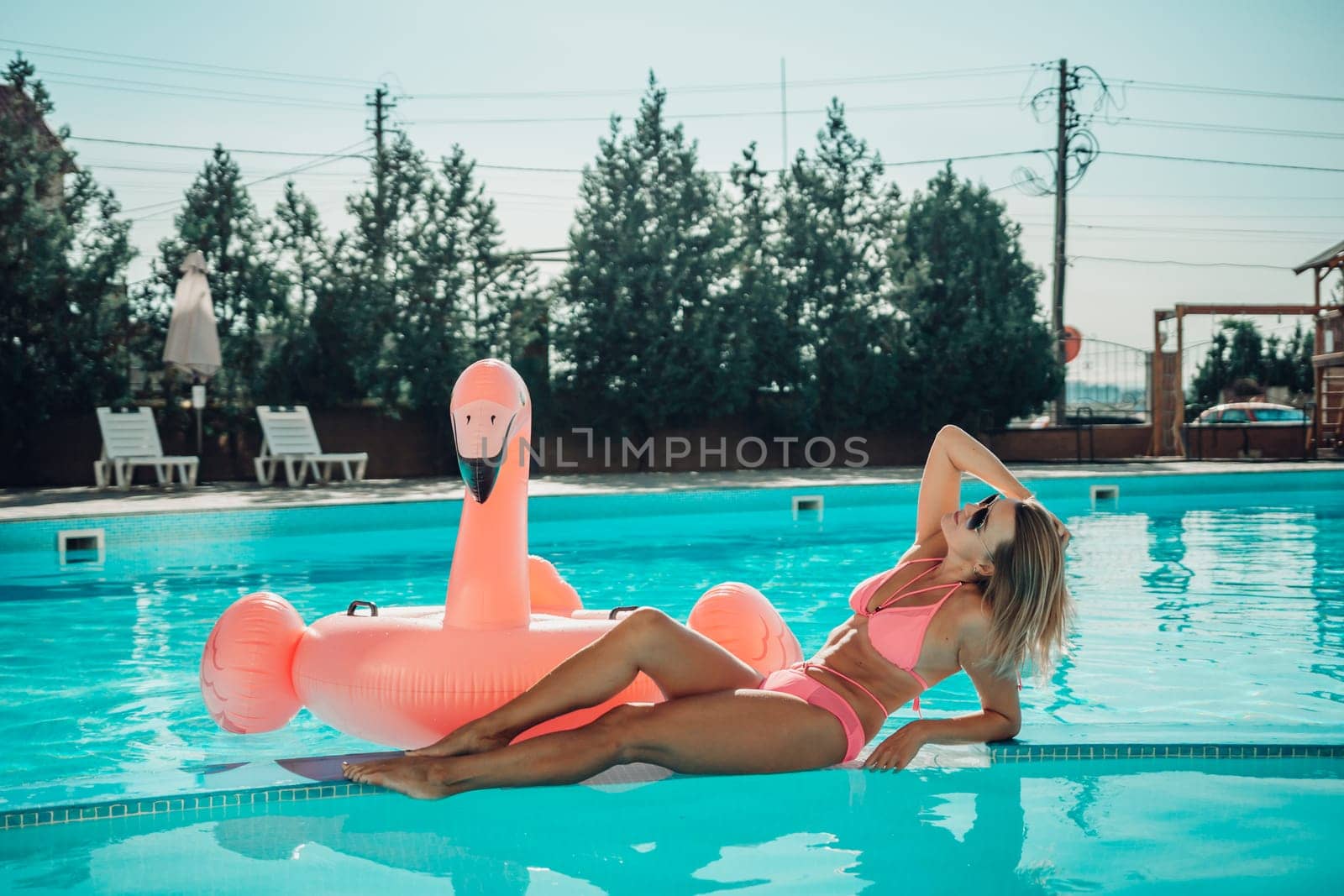A woman is laying on a pink inflatable flamingo in a pool. The scene is bright and cheerful, with the pink flamingo adding a playful touch to the image