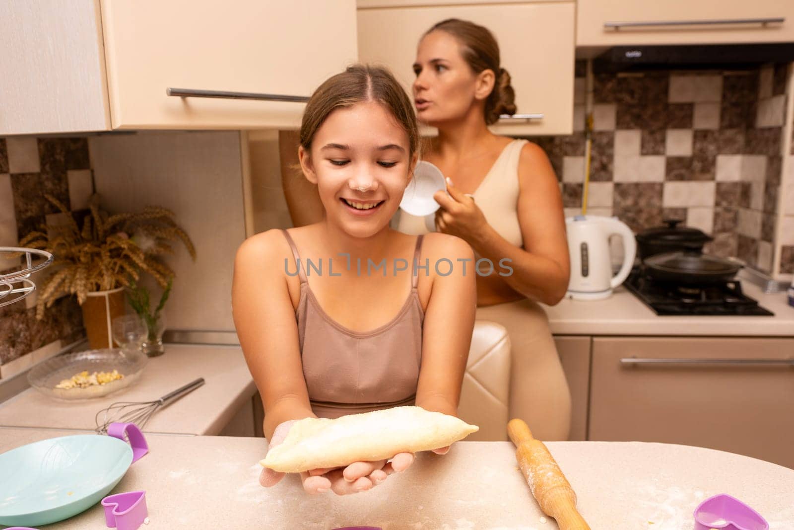 A young girl smiles as she holds up a ball of dough while her mother looks on in a kitchen.