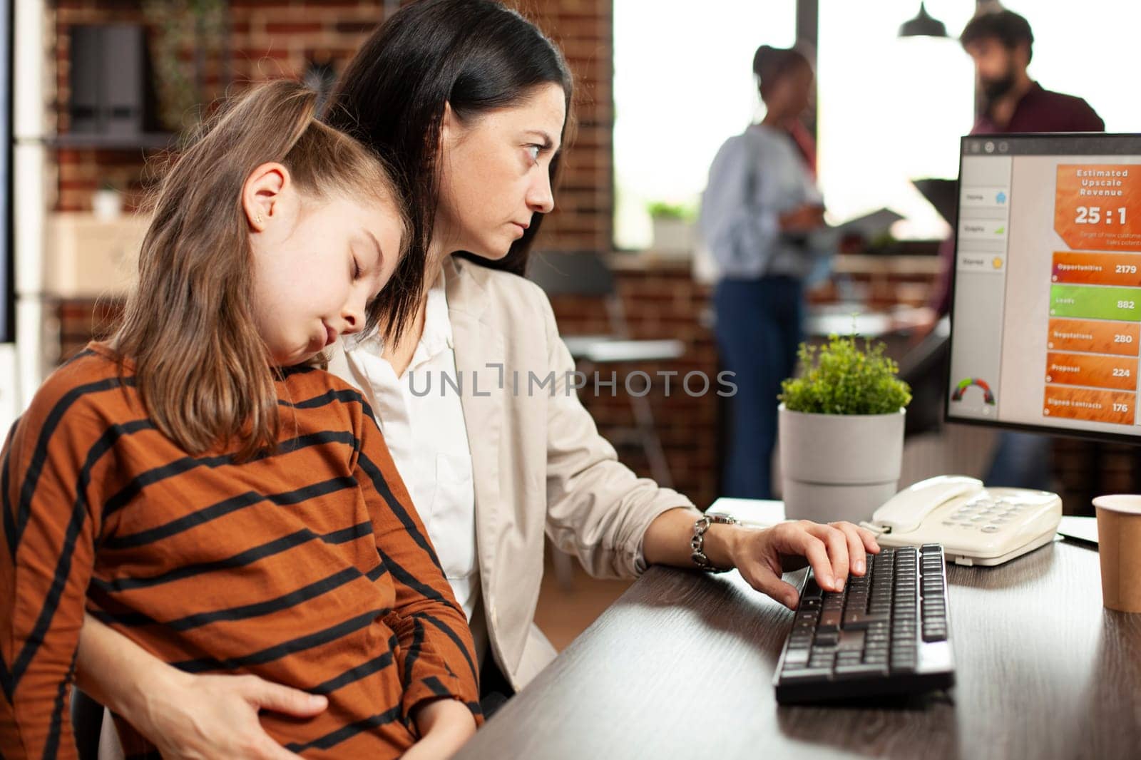 Working businesswoman holding her sleepy daughter at startup office desk, with desktop pc and telephone nearby. Young mother reviews market analysis on computer as little girl sleeps on her shoulder.