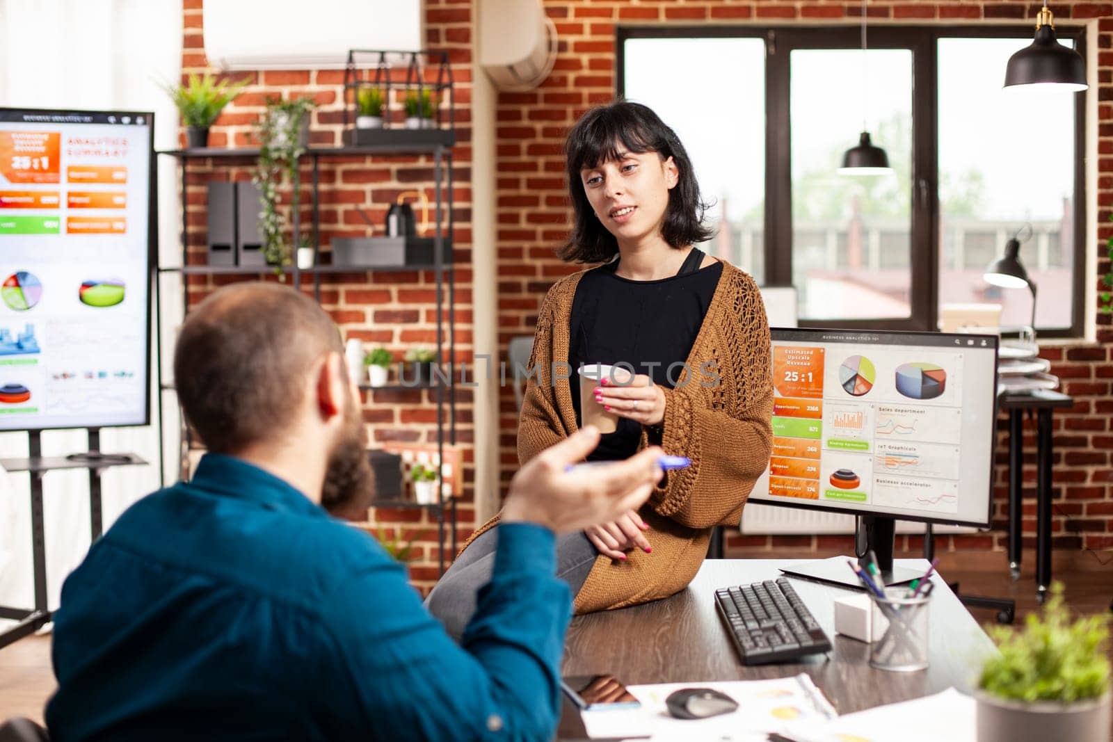 Female manager sitting table and holding coffee cup as employee explains marketing strategy for startup company. Businesswoman listening to young man describe financial analysis at brick wall office.