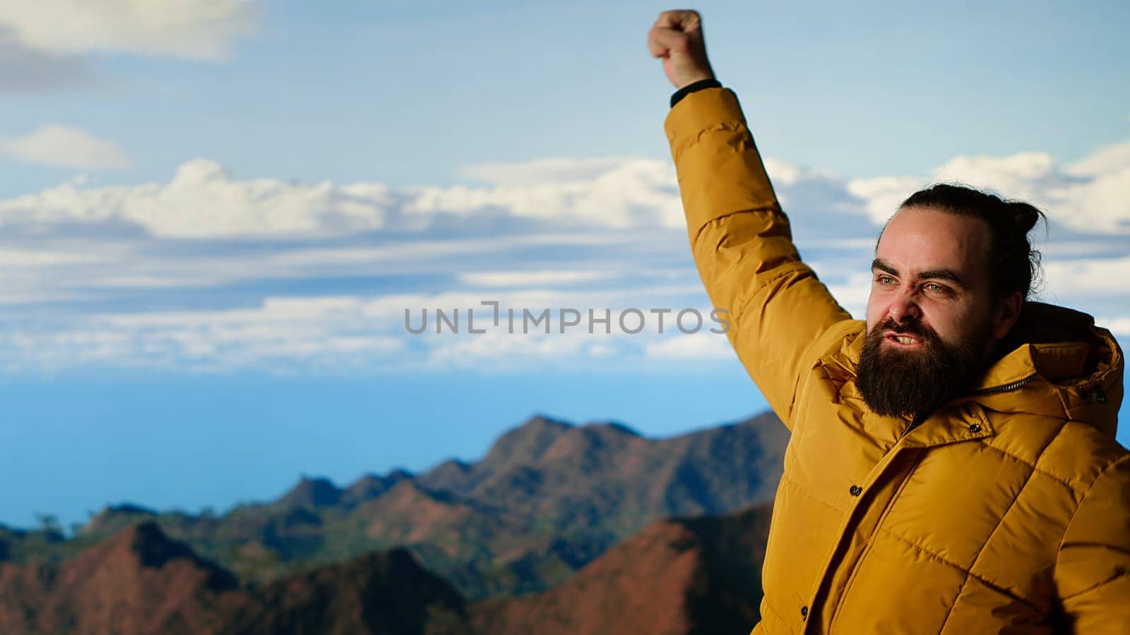 Triumphant hiker stands at the mountain peak with his arms raised in victory, celebrating success and personal achievement of climbing another mountain. Breathtaking panoramic view. Camera B.