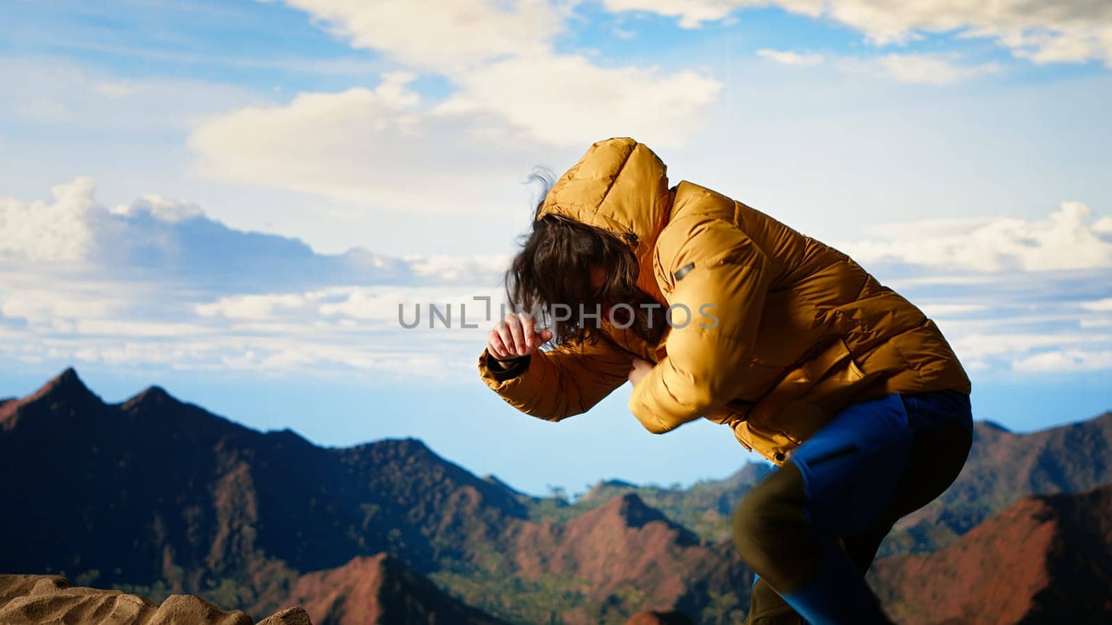 Clumsy traveler loses balance on a steep mountain peak as powerful winds knock him off course, falling and being unequipped in expedition. Unprepared hiker for the cold weather. Camera B.