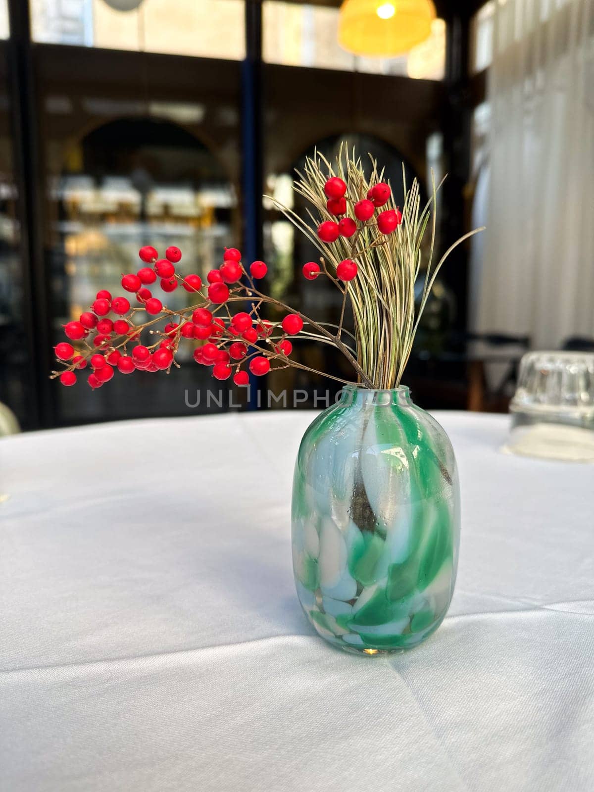 Decorative vase with red berries and pine branches on restaurant table. Minimalism, elegance, and seasonal interior decor in public spaces