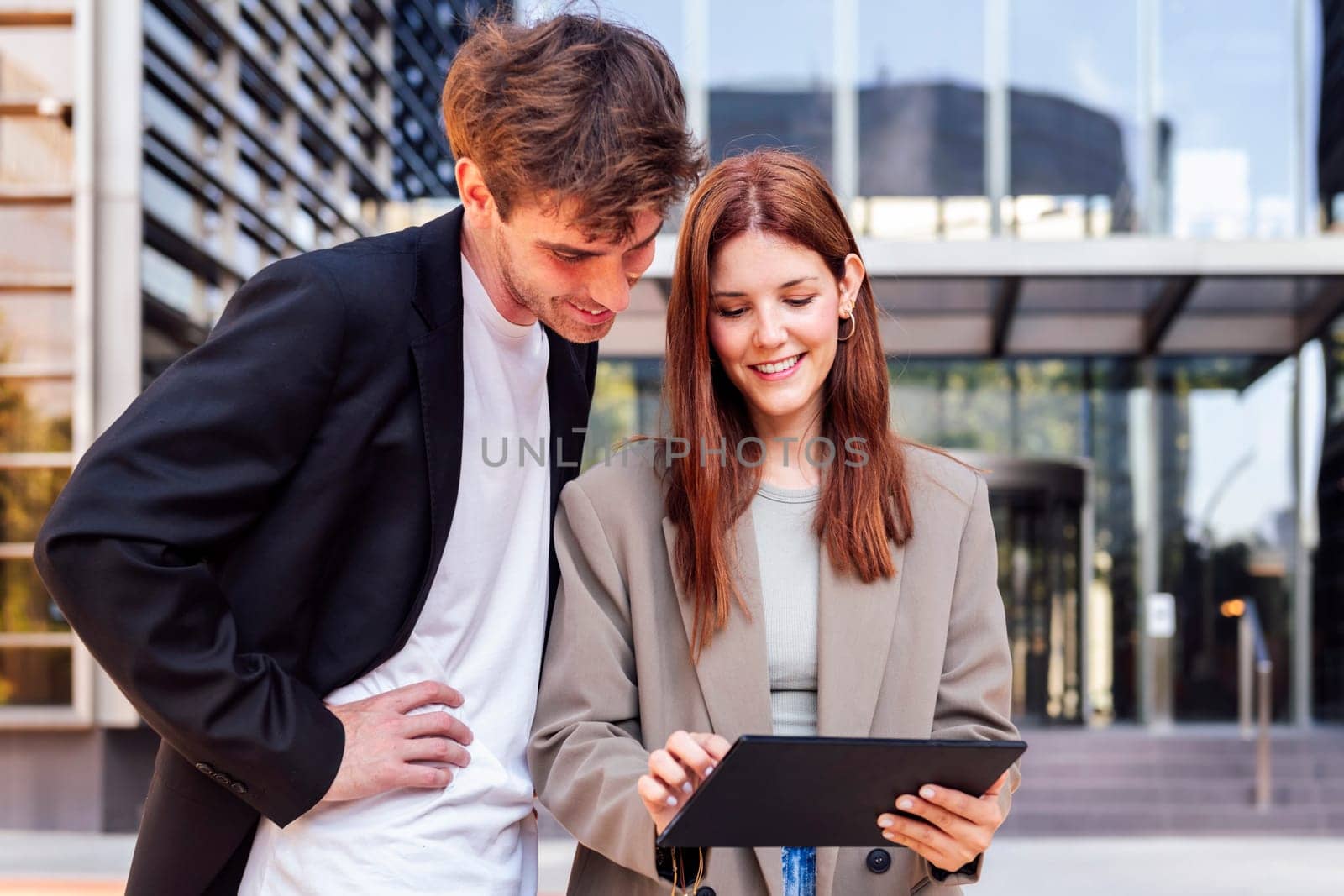 man and a woman standing outside in front of office building, the man is pointing at digital tablet in the woman hand, concept of business and technology