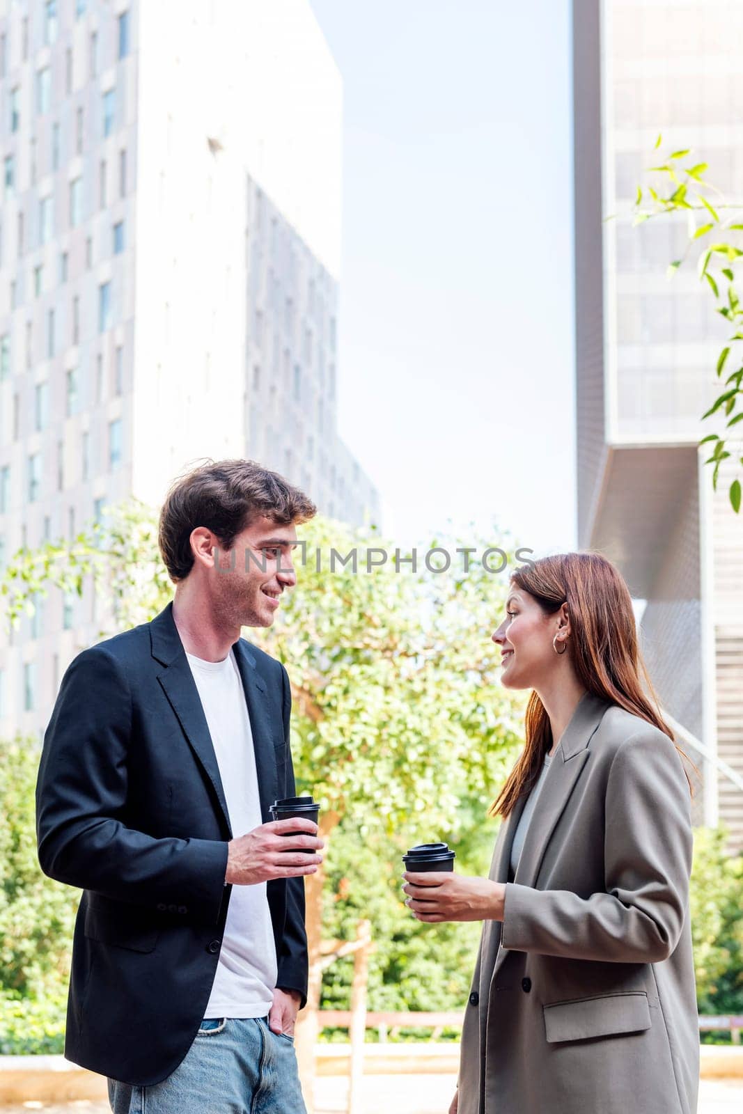 smiling man and a woman standing in a park near of office buildings talking while taking coffee, they are smiling and enjoying with the company, concept of business, copyspace for text