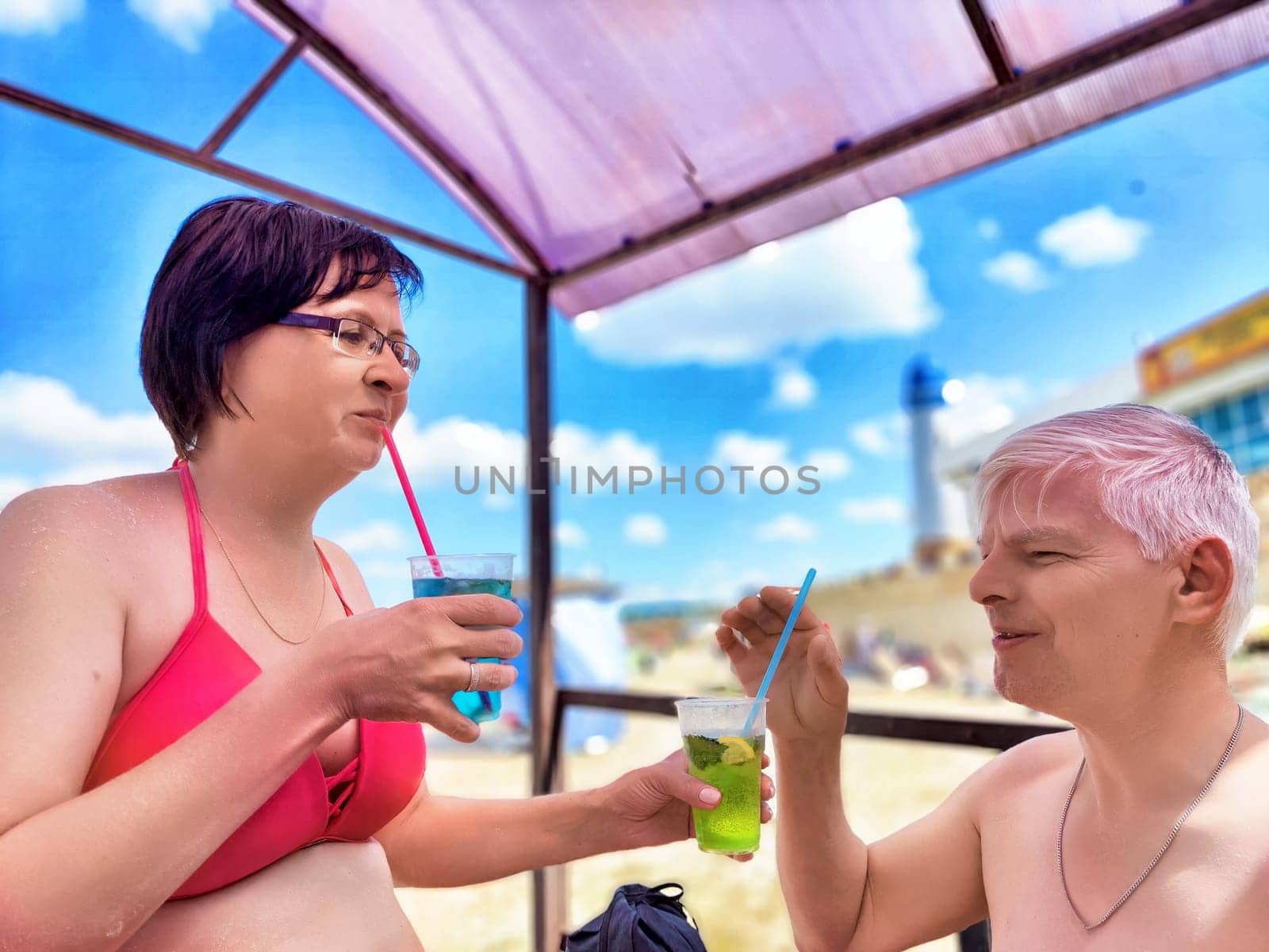 Middle-aged couple savoring cool beverages while relaxing under a cabana at the beach. They share smiles and laughter during their vacation in a warm coastal setting.