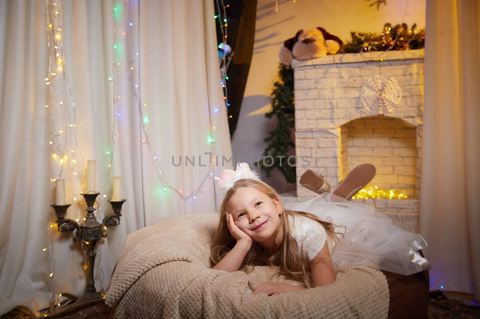 A happy child in a white dress enjoys a peaceful moment in a warmly decorated room filled with Christmas lights and holiday cheer.